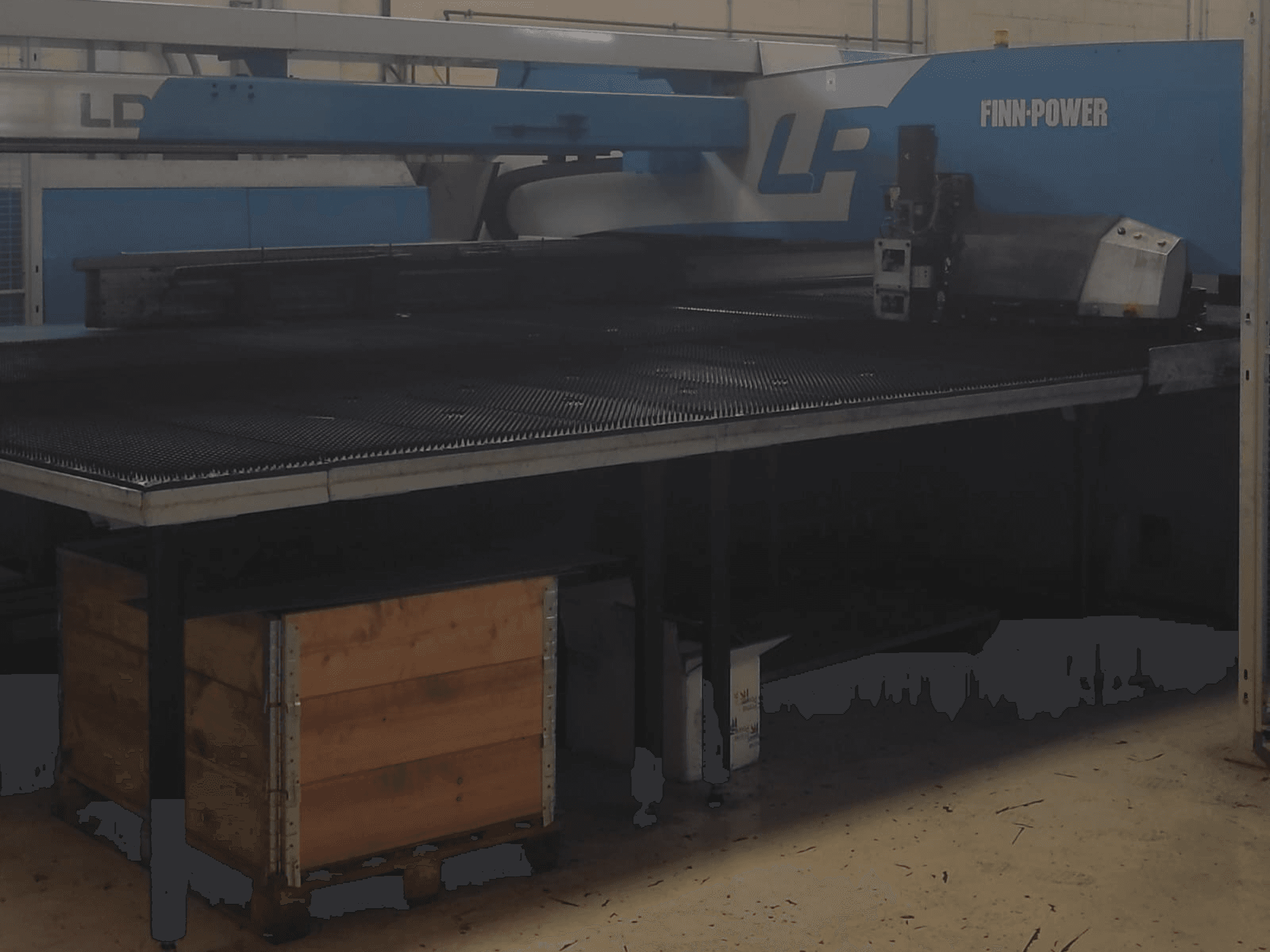 Режущий станок Finn-Power LP на заводе, вид спереди с текстурированным металлическим столом и деревянным ящиком под ним.