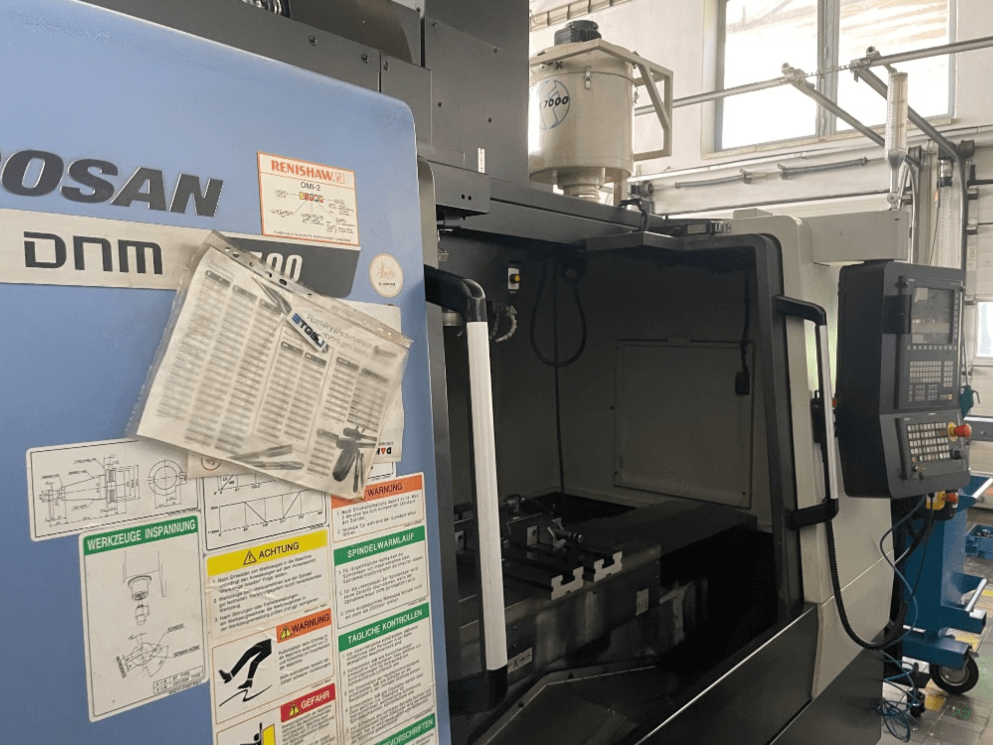 Вертикальный обрабатывающий центр DOOSAN DNM 5700, вид спереди, показывает панель управления, знаки безопасности и детали станка.