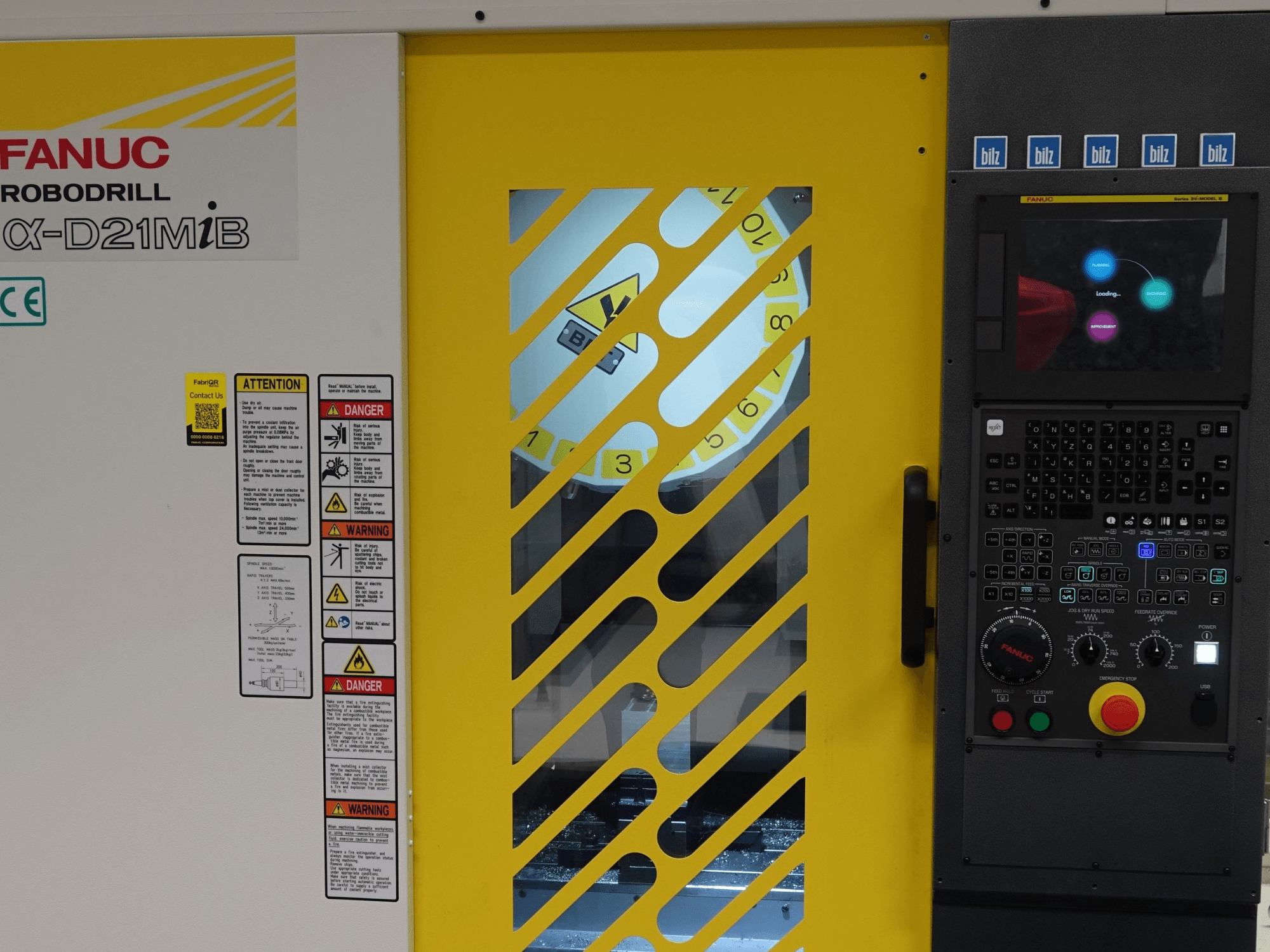 Вертикальный обрабатывающий центр FANUC α-D21MiB, вид спереди, с указателями безопасности, панелью управления и желтой защитной дверцей.