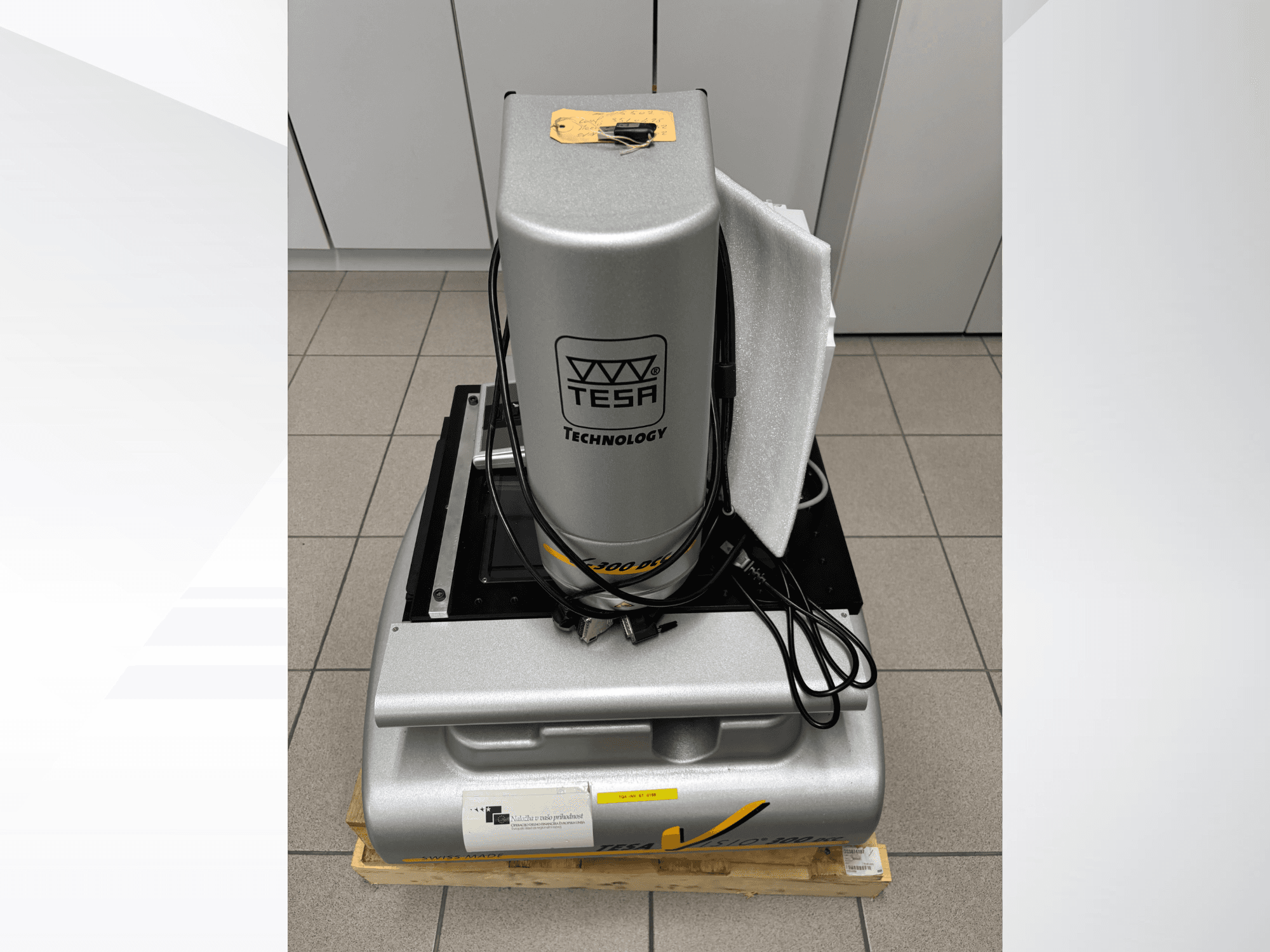 Оптическая измерительная машина TESA Visio 300 DCC на переднем плане с кабелями, на деревянном поддоне, видна защитная упаковка.