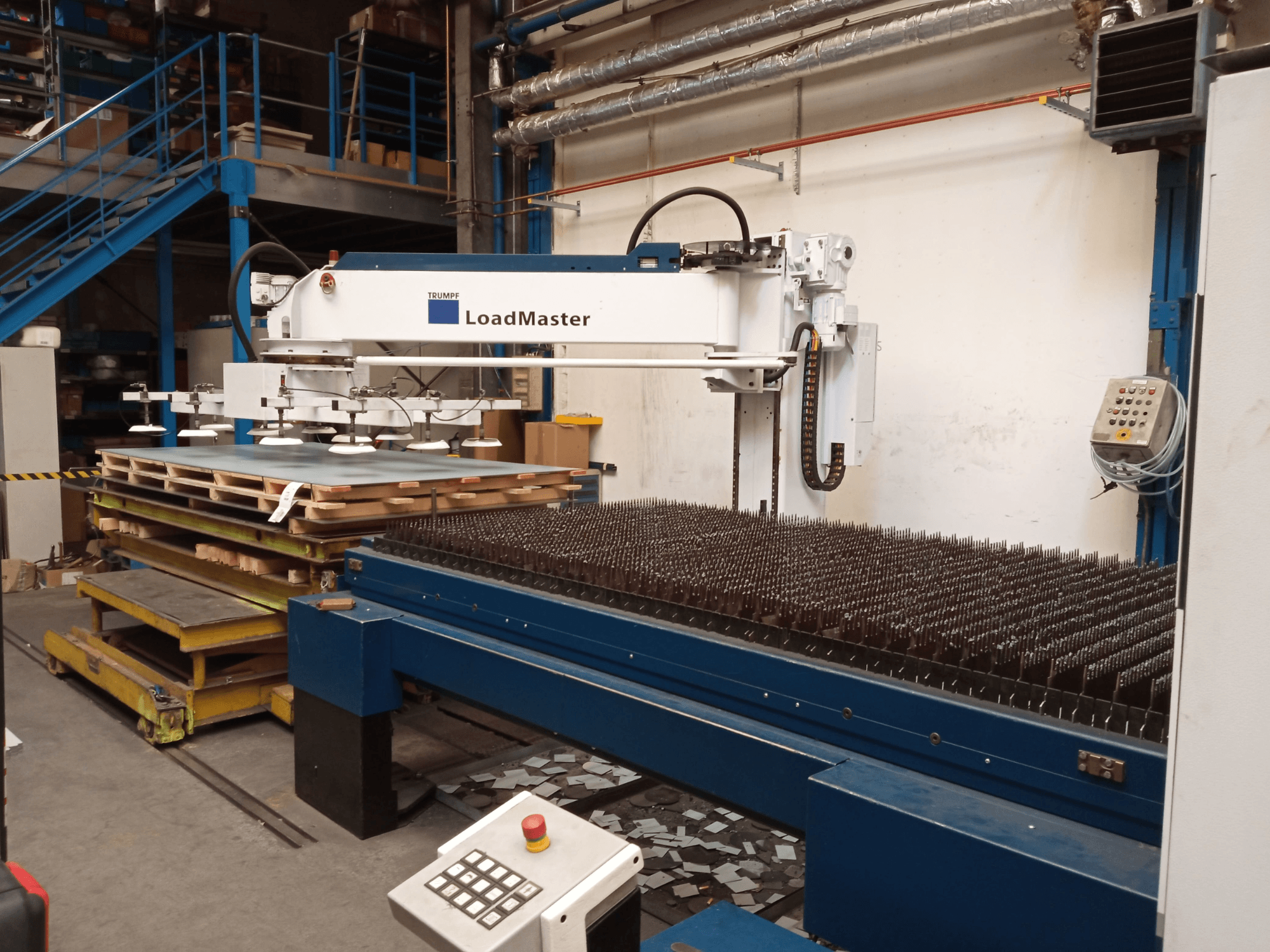 Лазерный резак TRUMPF TruLaser 3030 с CO₂-лазером в виде сбоку, оснащенный системой LoadMaster и поддонами с материалами.