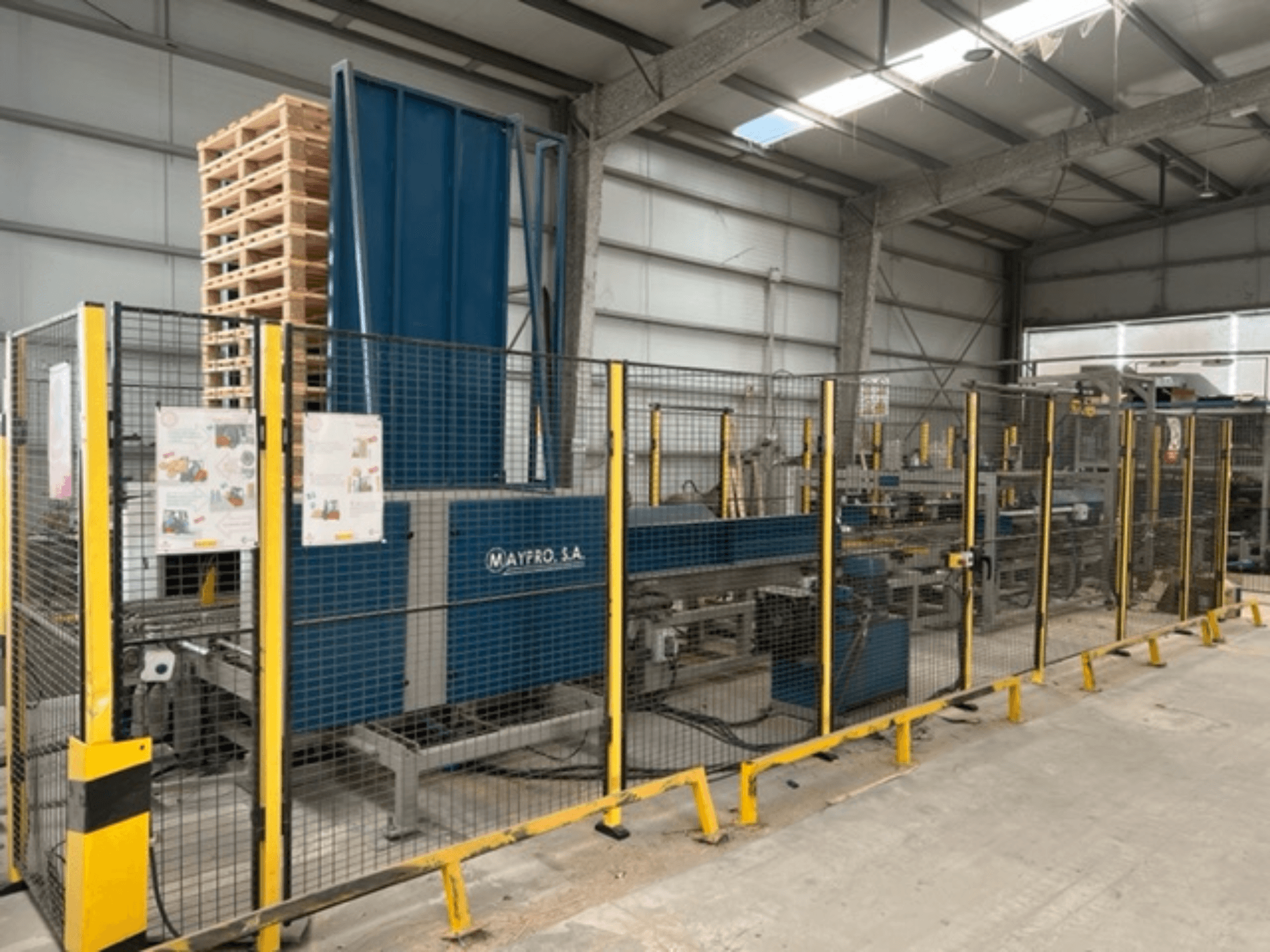 Промышленная машина для упаковки паллет производства Maypro S.A. на складе, с защитной сеткой и элементами управления, вид сбоку.