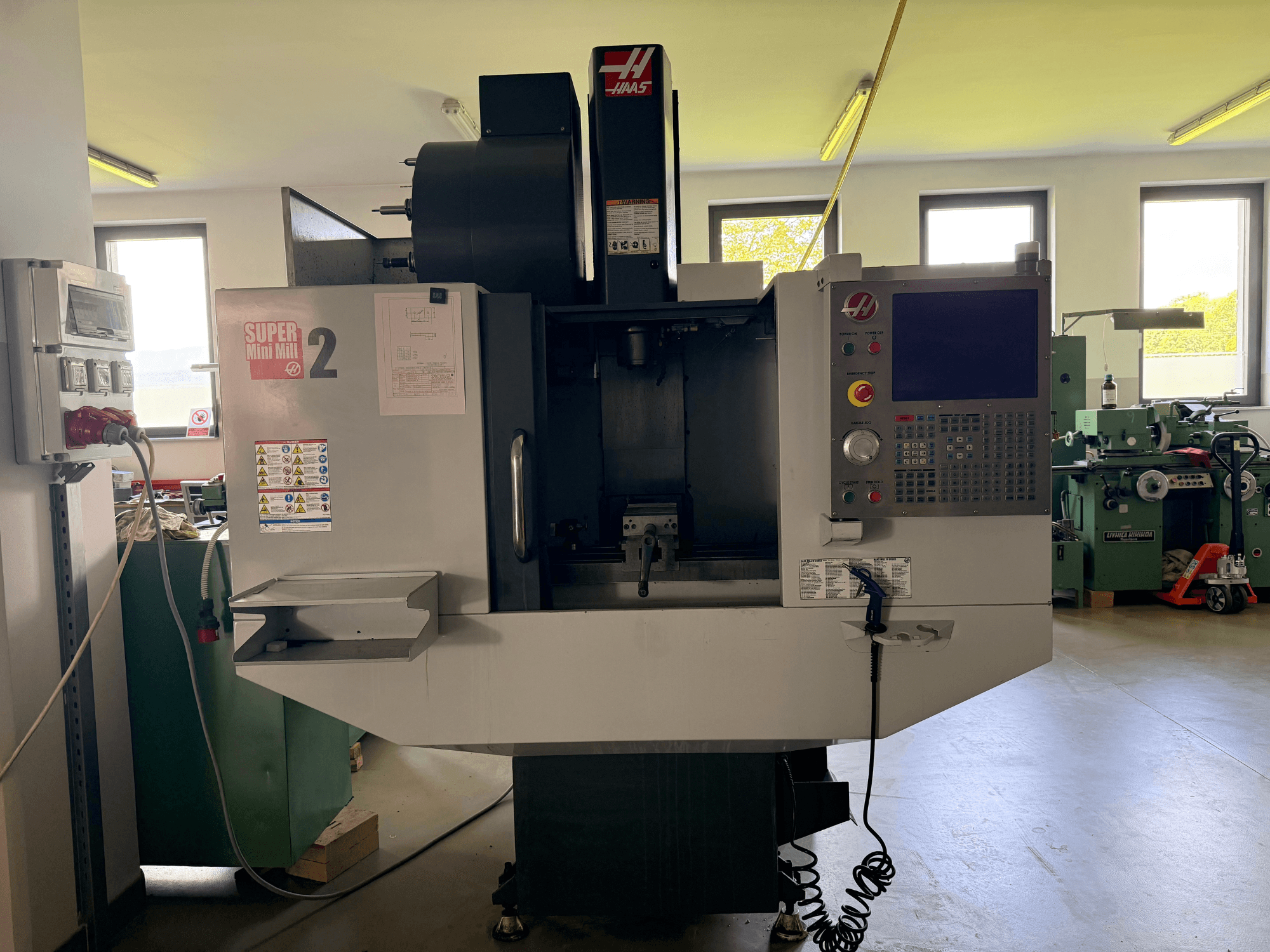Вертикальный обрабатывающий центр Haas Mini Mill 2 на переднем плане, панель управления и зона инструментов с предупреждающими надписями.