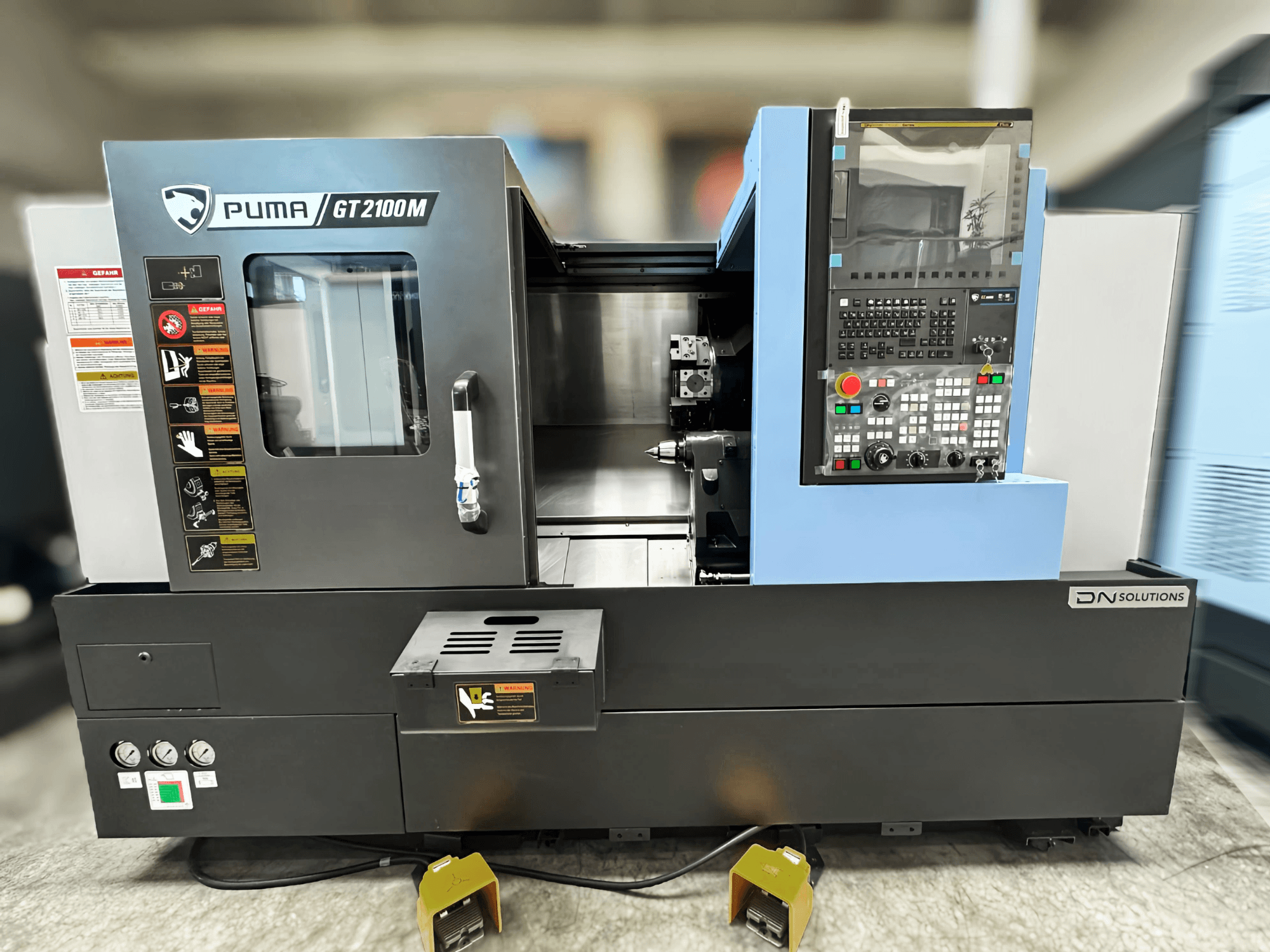 Токарно-фрезерный центр DN SOLUTIONS PUMA GT2100M, вид спереди, демонстрирующий панель управления, камеру обработки и знаки безопасности.