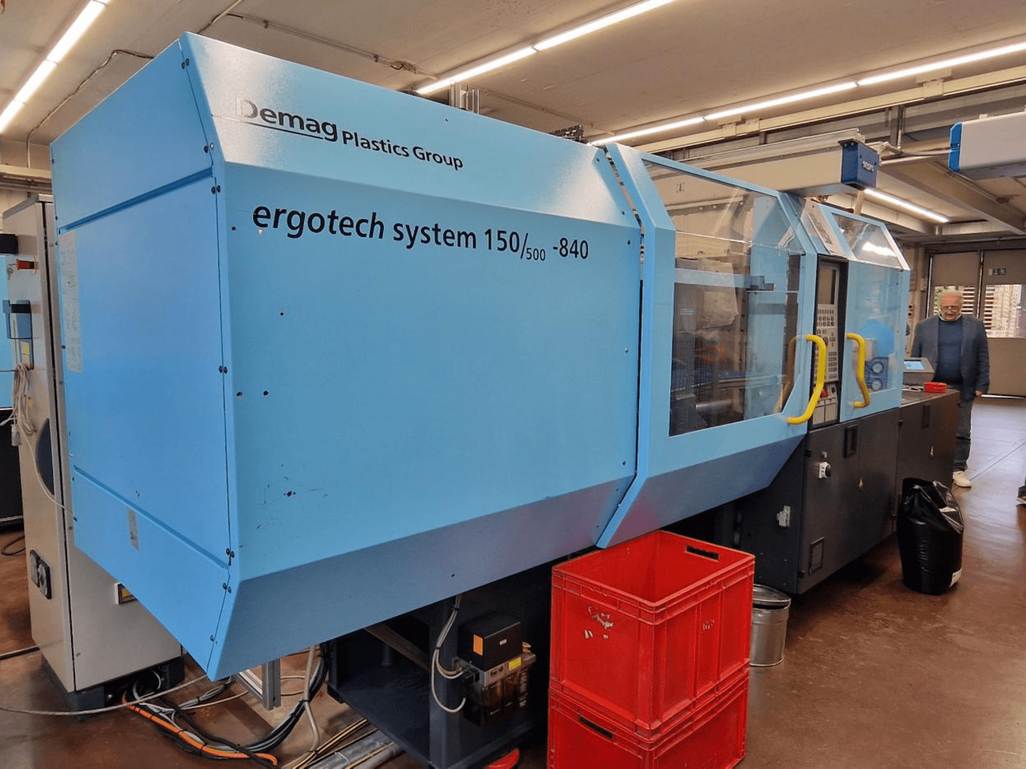 Термопластавтомат Demag Ergotech System 150/500-840 синего цвета, вид сбоку, с ящиками и оператором на заднем плане.