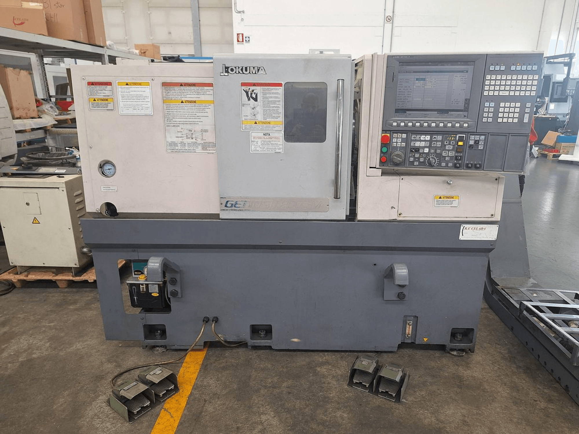 Горизонтальный токарный станок Okuma GENOS L200E-M вид сбоку; панель управления, предупреждения о безопасности и ножные педали.