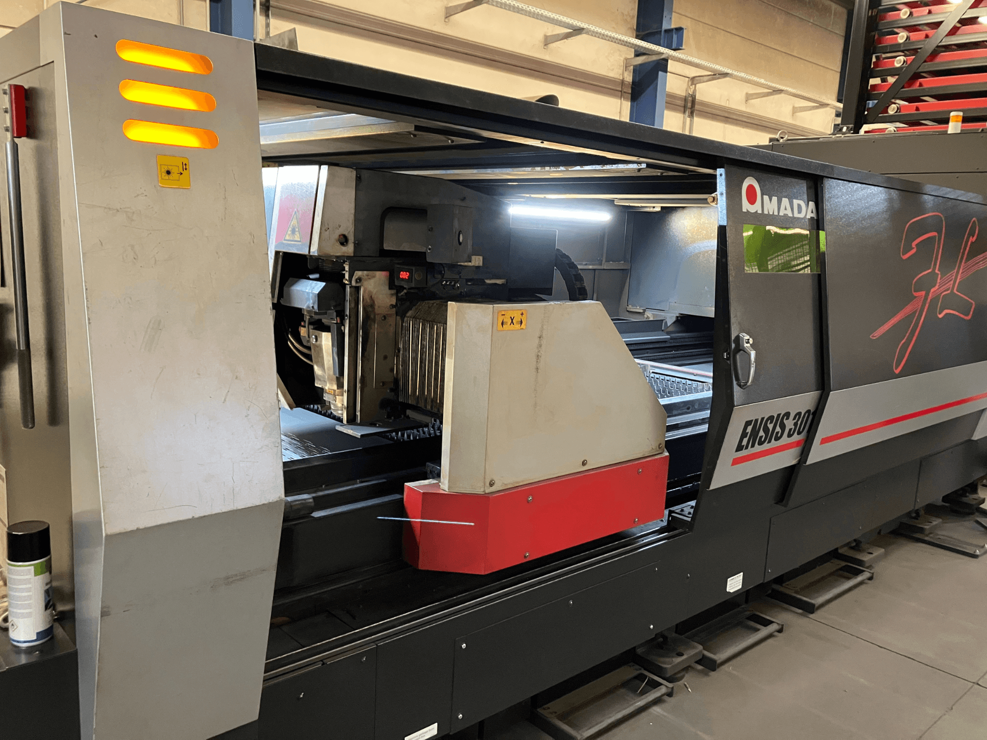 Станок волоконно-лазерной резки AMADA ENSIS 3015 AJ, вид спереди, изображение режущей головки и органов управления внутри металлической рамы.