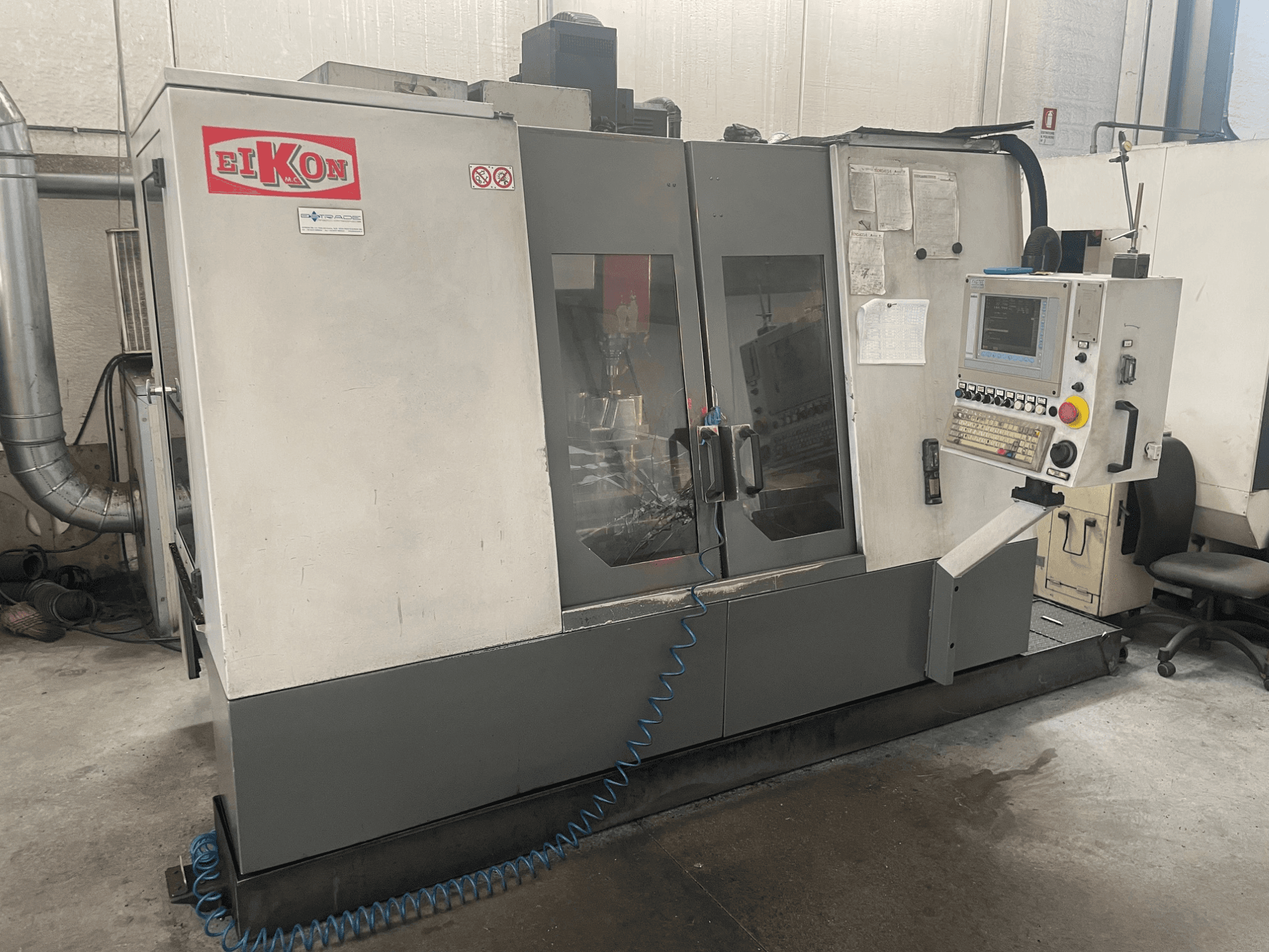 Станок с ЧПУ EIKON с видом спереди: панель управления, большие стеклянные двери и синий воздушный шланг на полу.