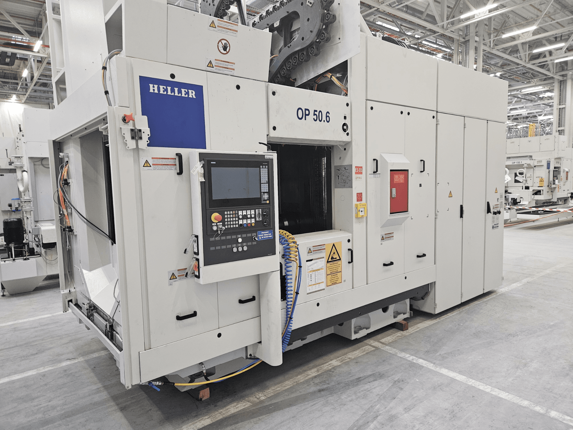 Обрабатывающий центр Heller OP 50.6, вид спереди, с панелью управления, предупреждающими надписями и шлангами, видимыми в заводских условиях.
