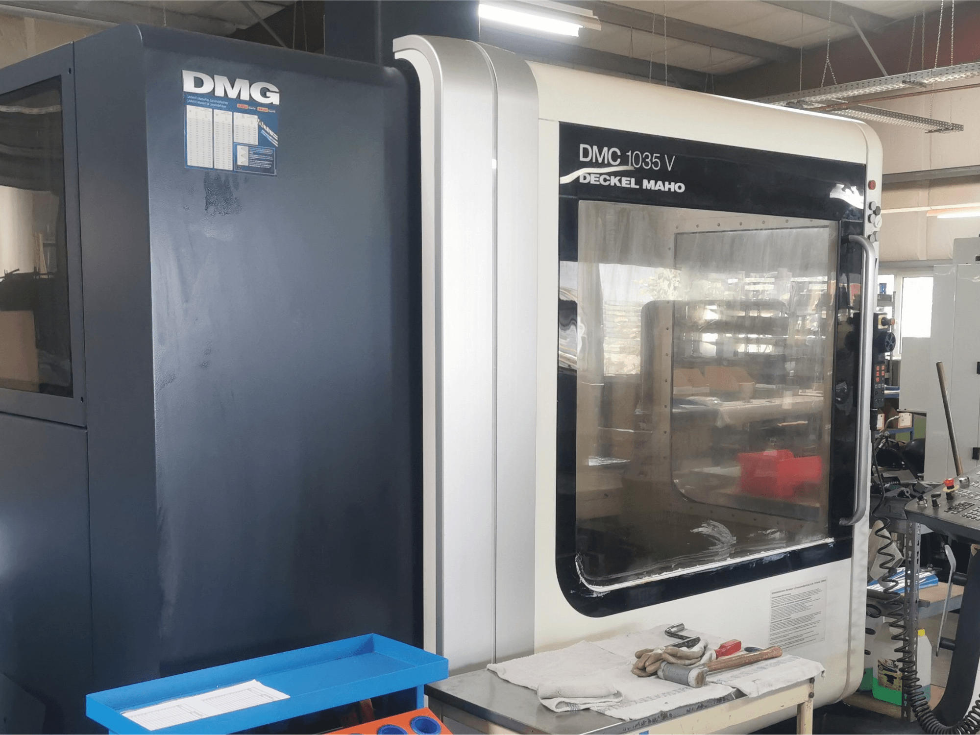 Фрезерный станок DMG DMC 1035 V в мастерской, вид спереди с синим лотком для инструментов и перчатками на поверхности.