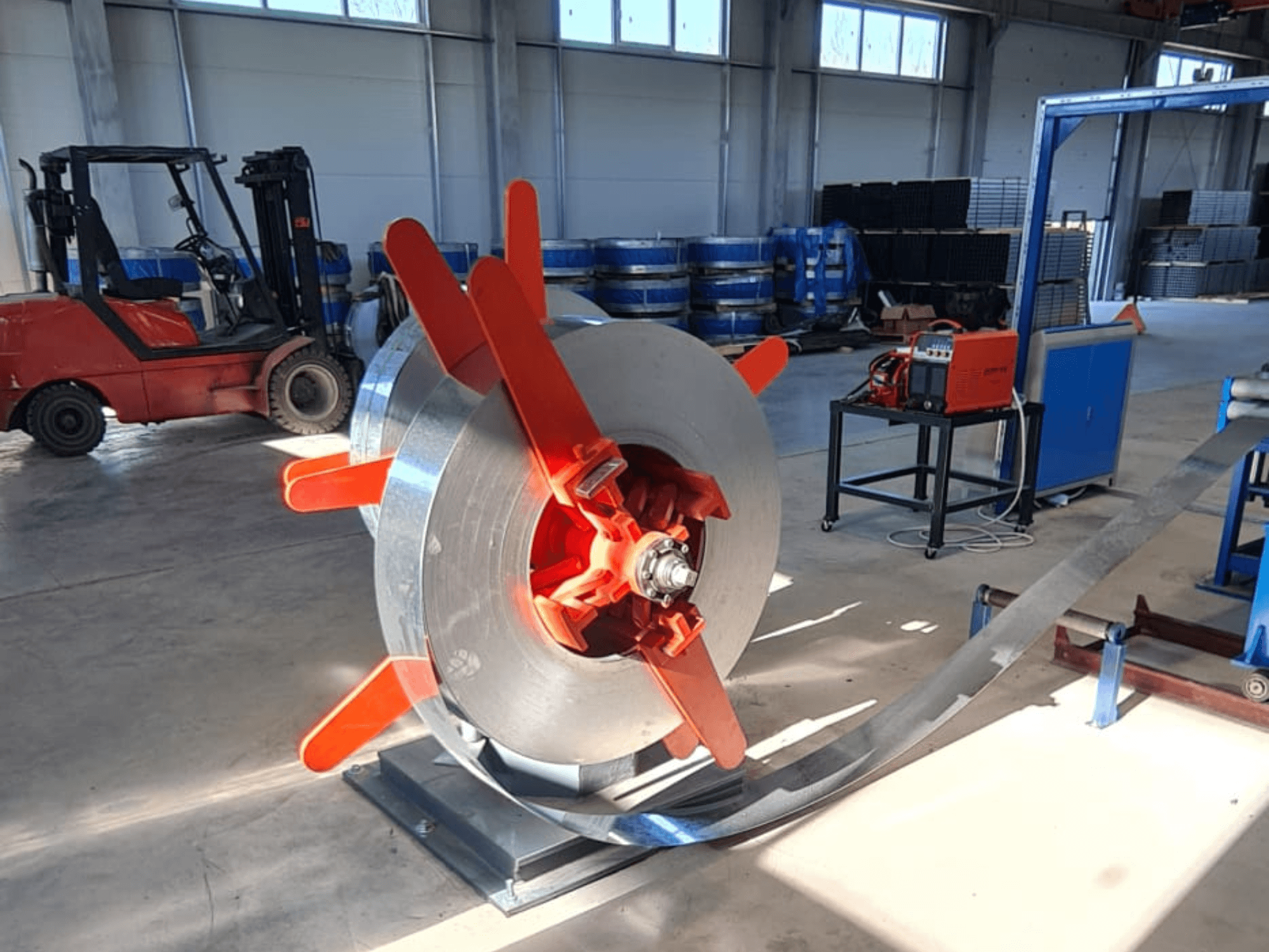 Металлическая катушка с красными ручками на подставке, вид под углом. Промышленный фон со станками и вилочным погрузчиком.