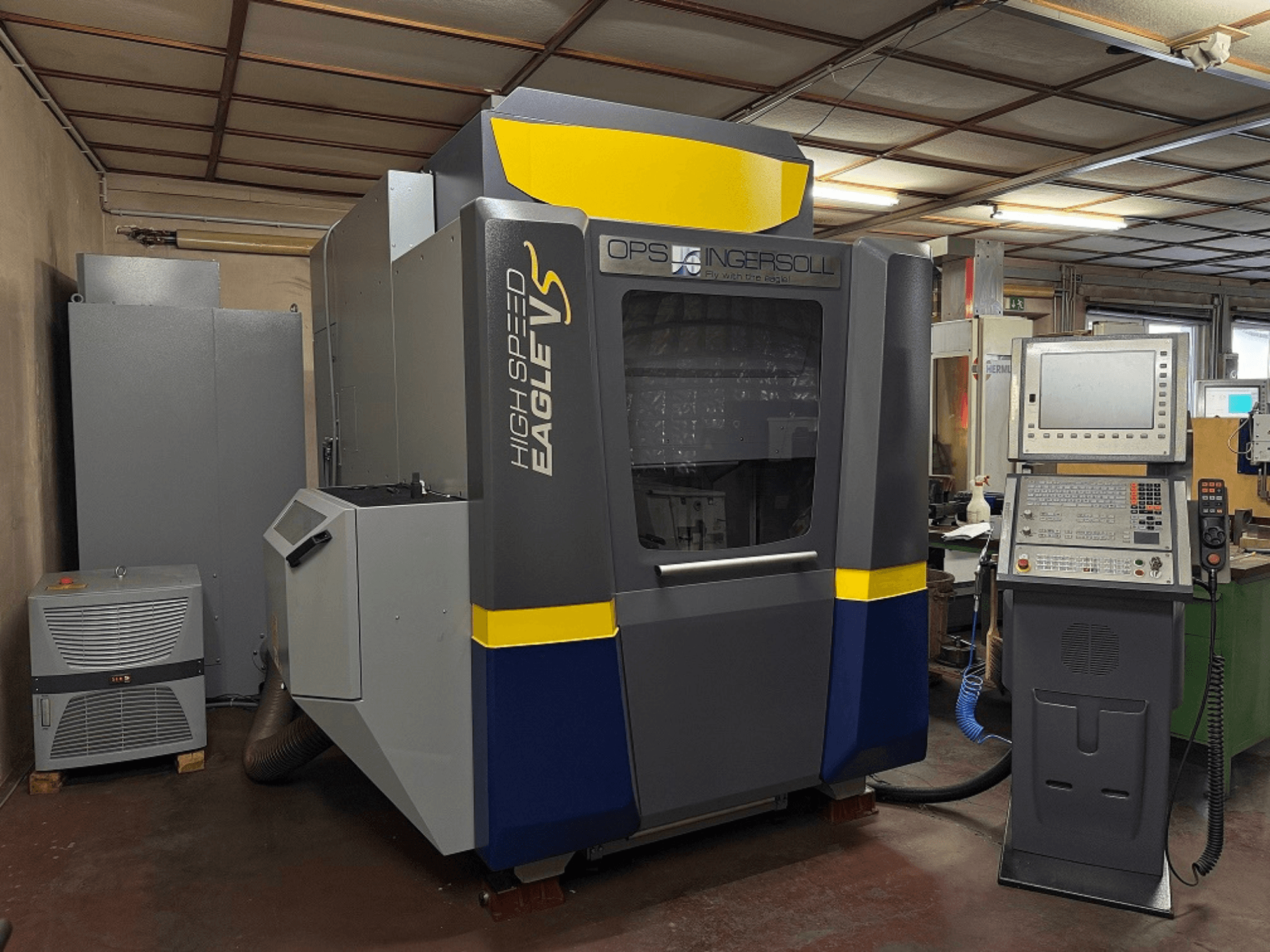 Высокоскоростной станок Eagle V производства OPS Ingersoll, вид сбоку, с панелью управления и соседним оборудованием в промышленной обстановке.