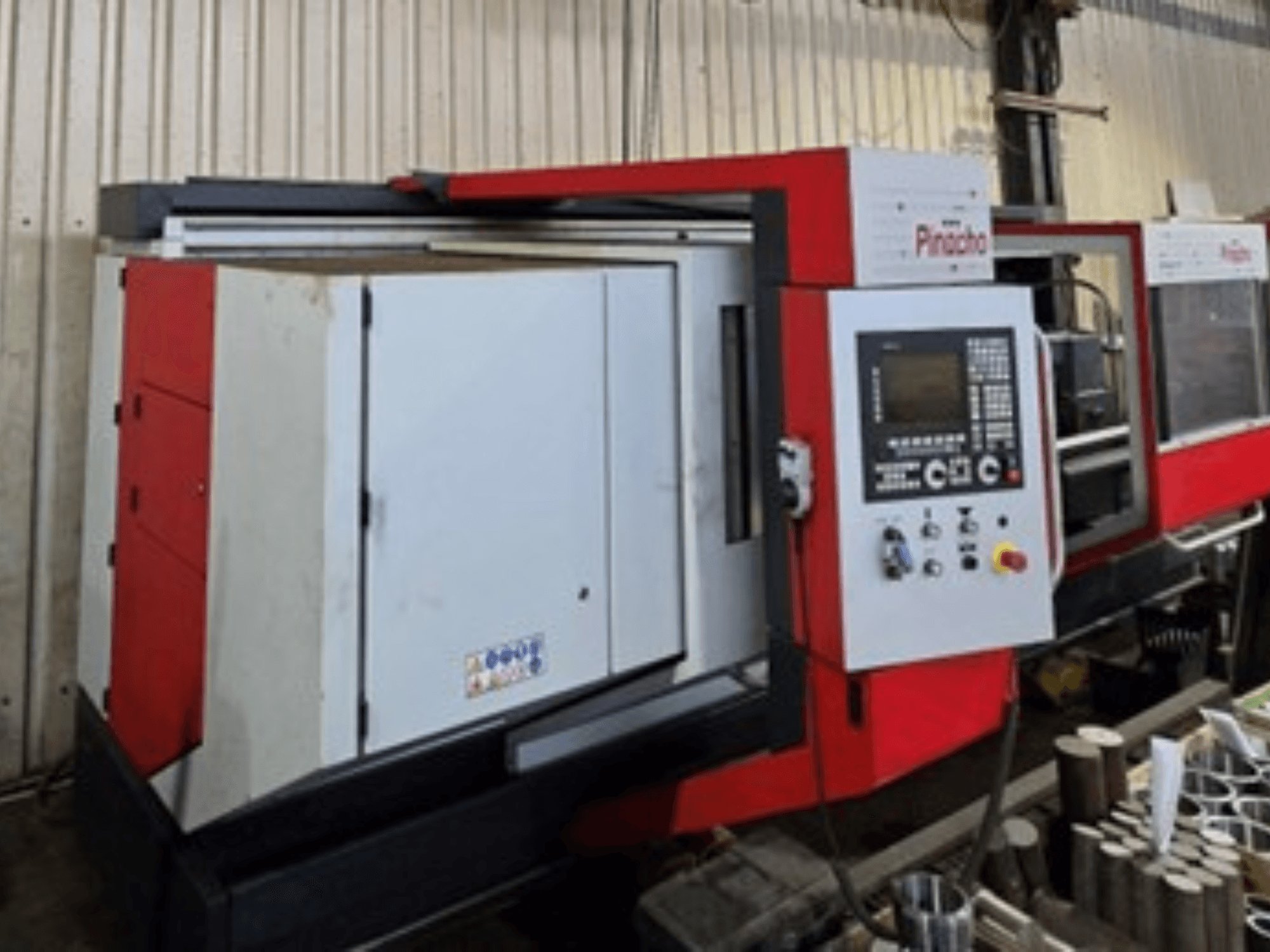 Красно-белый станок с ЧПУ Pinacho, вид сбоку, с панелью управления и материалами на полу, демонстрирующий оперативную настройку.