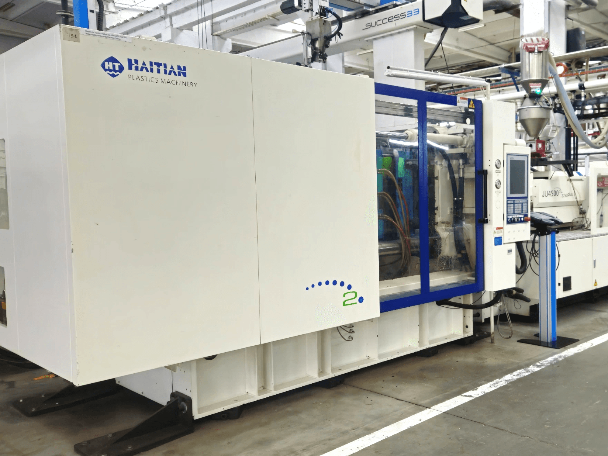 Модель Haitian Plastics Machinery, вид спереди, демонстрирующая оборудование с панелью управления и видимыми трубками, в промышленной обстановке.