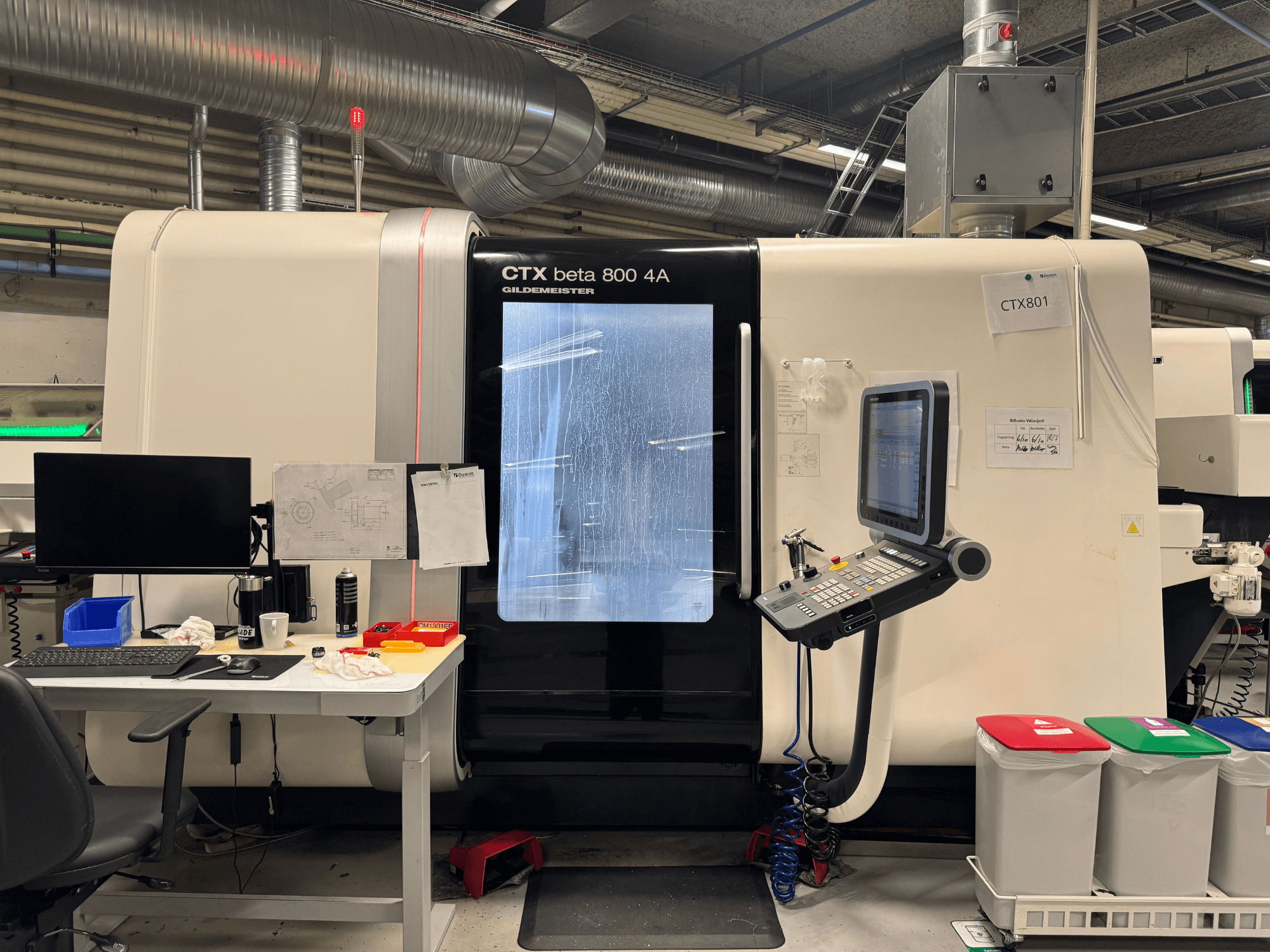 Машина CTX beta 800 4A с панелью управления и монитором, вид сбоку, в мастерской с офисным оборудованием и цветными мусорными контейнерами.