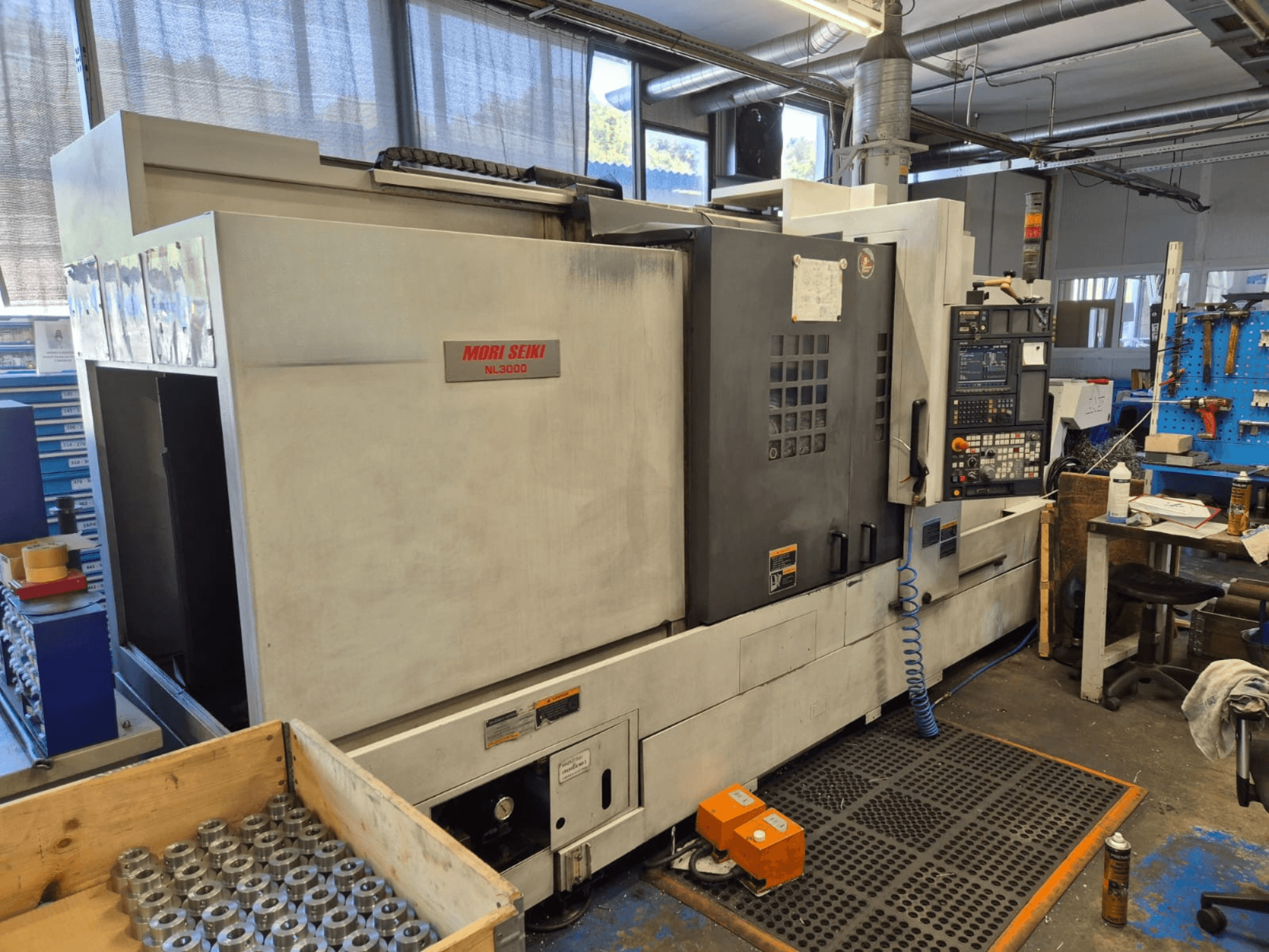Токарный станок с ЧПУ Mori Seiki NL3000 в мастерской, вид сбоку, демонстрирующий панель управления, инструменты и металлические детали в деревянном ящике.