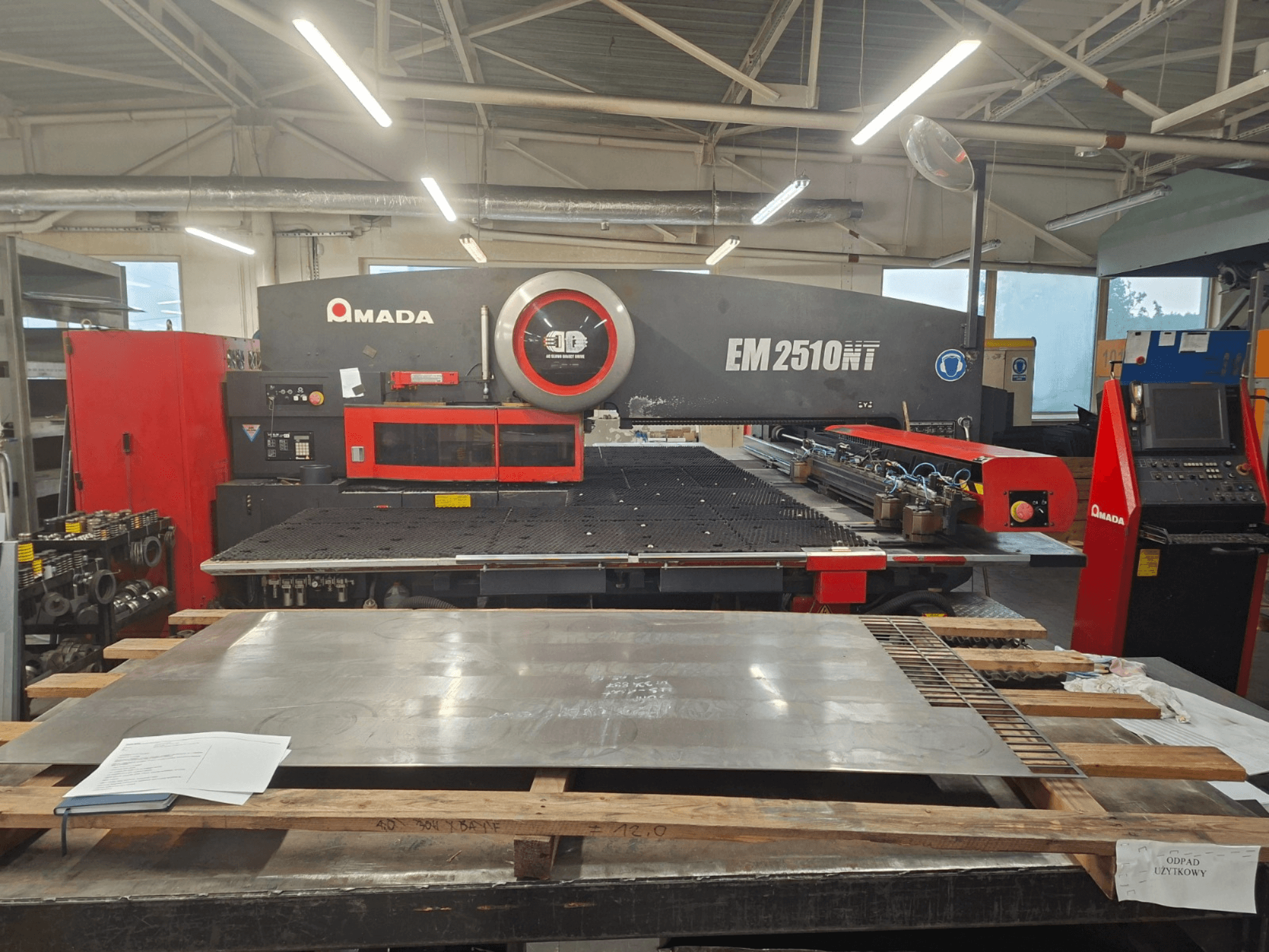 Вид спереди на револьверный вырубной пресс AMADA EM 2510NT, металлический лист и панель управления видны в мастерской.