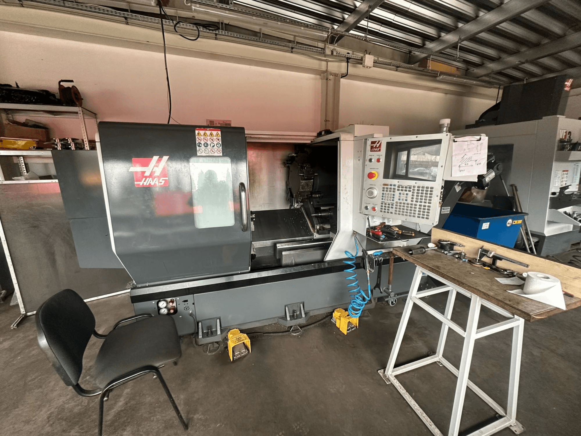 Токарный станок с ЧПУ Haas (модель ST-10) в мастерской, вид сбоку с изображением органов управления, инструментов на верстаке и стула рядом.