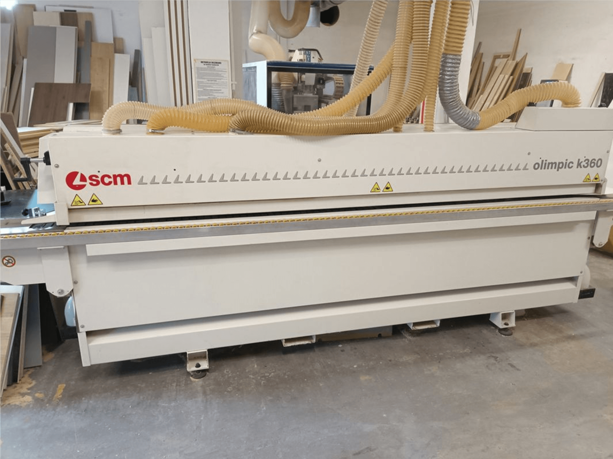 Кромкооблицовочная машина SCM Olimpic K360 T-ER1 в цехе, вид спереди с пылеотводящими шлангами и предупреждающими надписями.