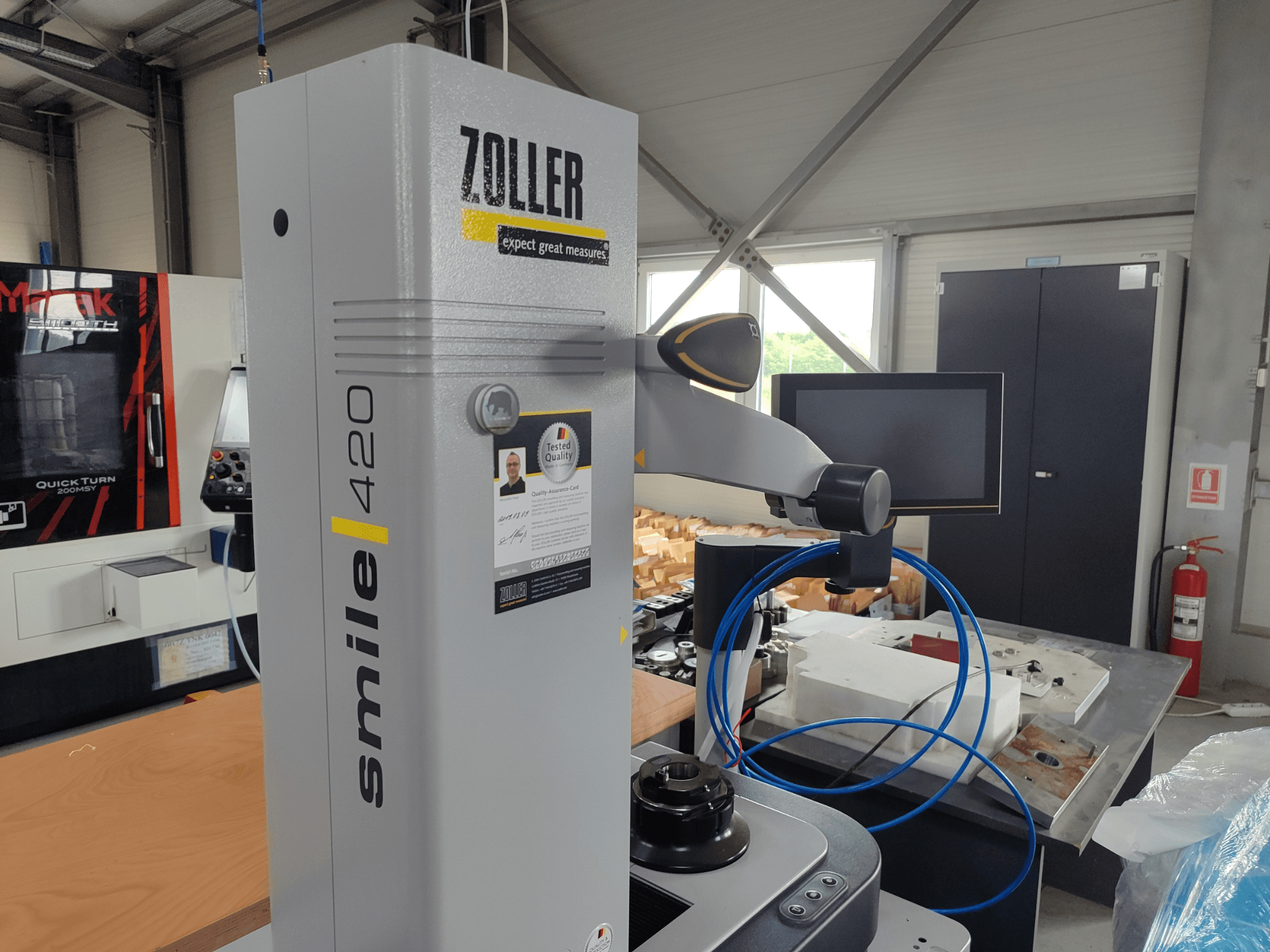 Измерительный прибор ZOLLER smile 420 с монитором, вид сбоку в мастерской, с синими шлангами и механизмами на заднем плане.