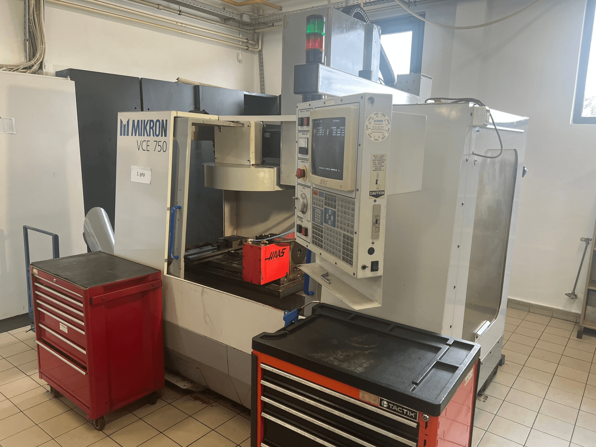 Alt text: "Вертикальный обрабатывающий центр MIKRON VCE 750, вид спереди с панелью управления и красными ящиками для инструментов, в просторной мастерской".