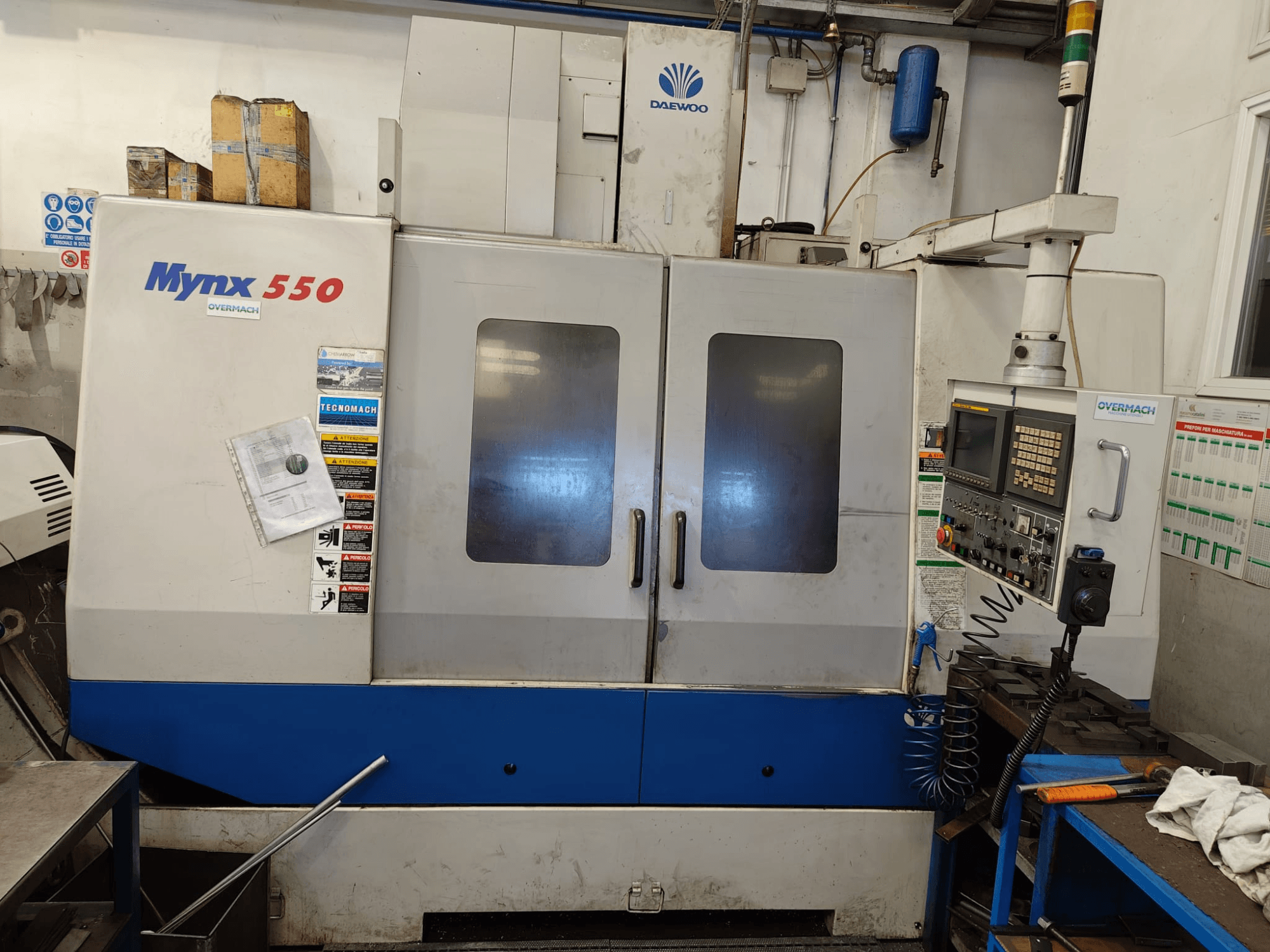 Вертикальный обрабатывающий центр DAEWOO MYNX 550, вид спереди, с панелью управления, двумя большими дверцами и предупреждающими надписями.