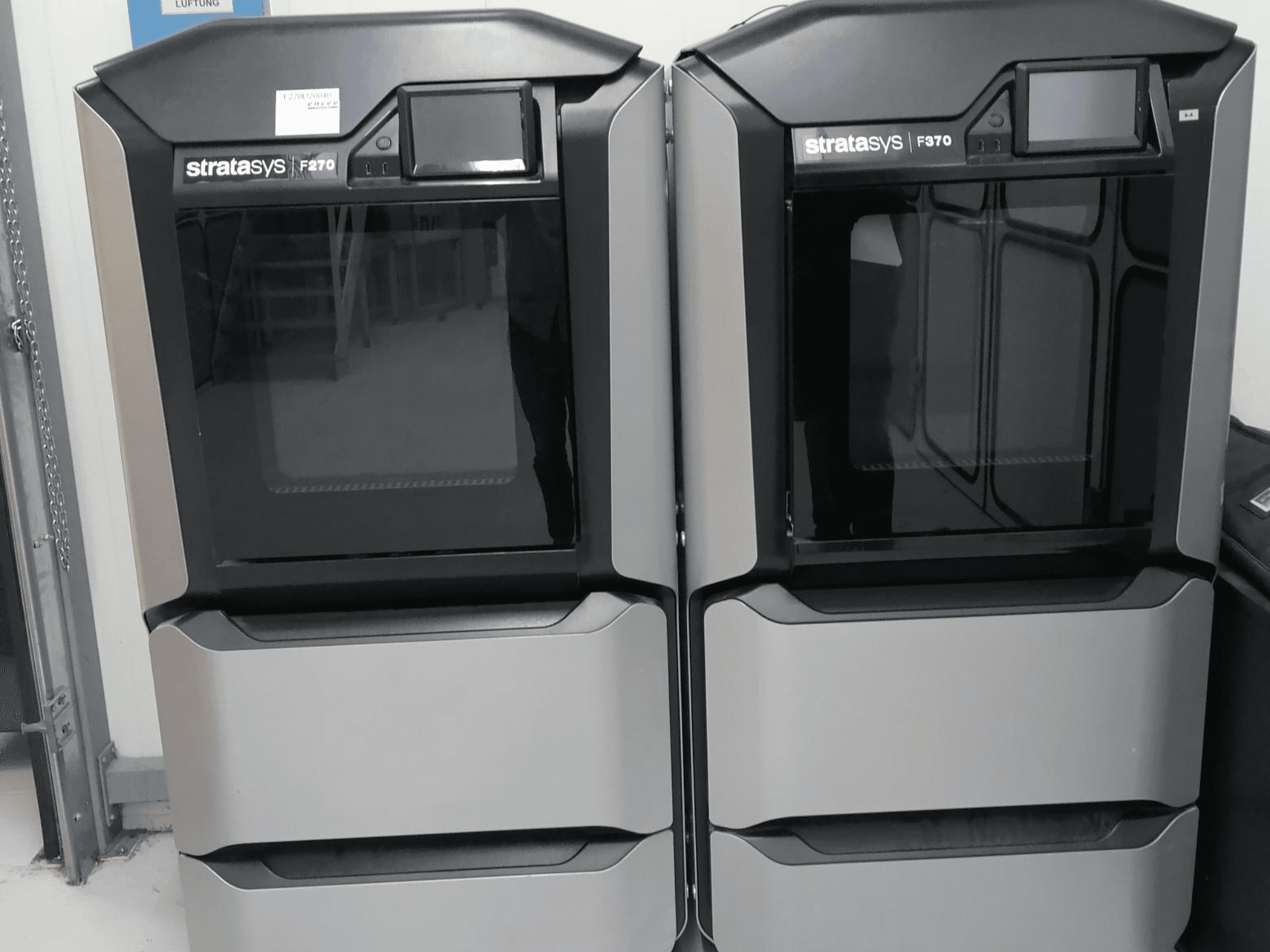 Пластиковый 3D-принтер Stratasys F270 с видом спереди: сдвоенные машины с гладкими серыми внешними поверхностями и цифровыми дисплеями.