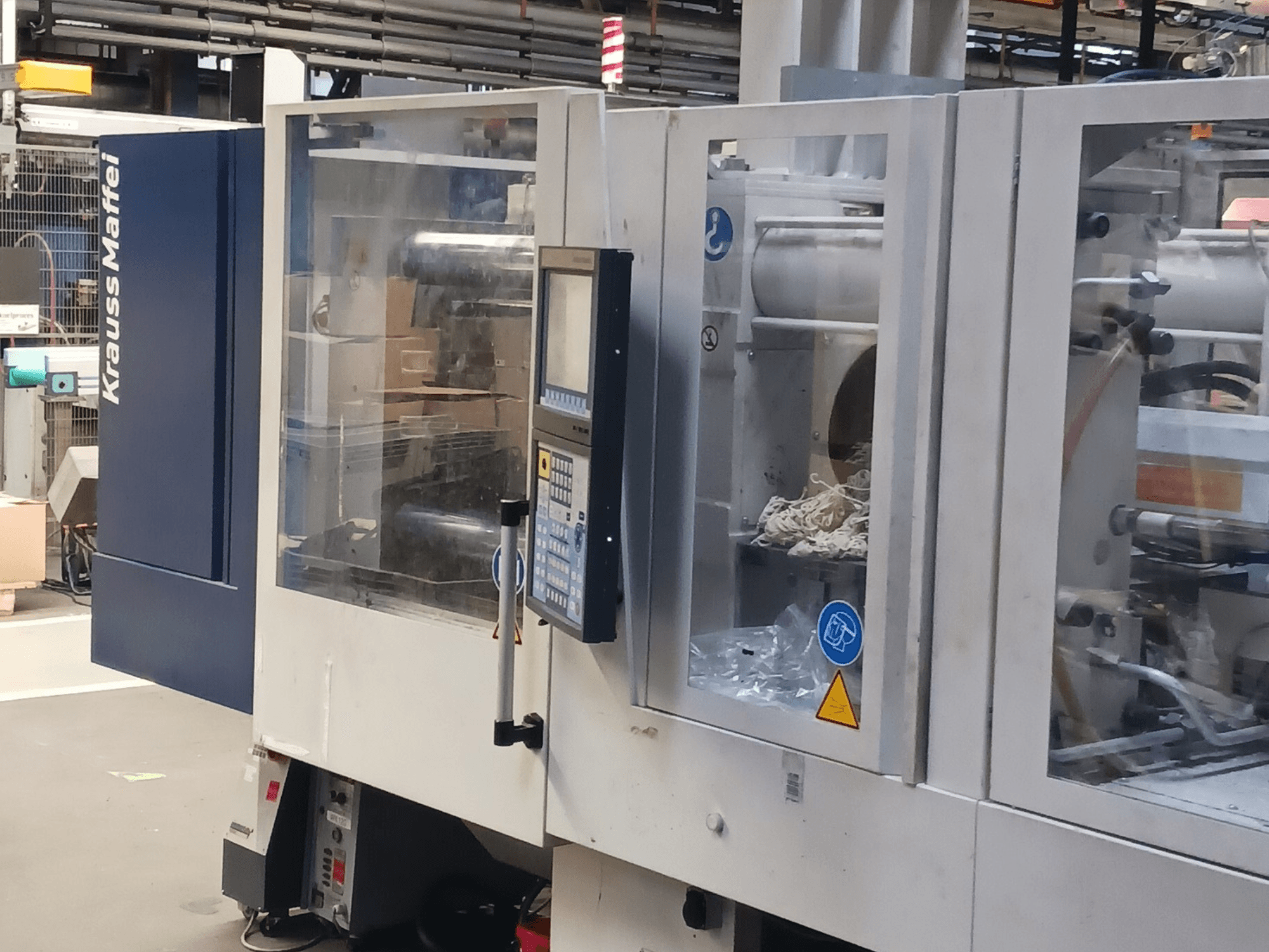 Термопластавтомат KraussMaffei, вид сбоку, на фоне панели управления, механизмов и упаковочных материалов.