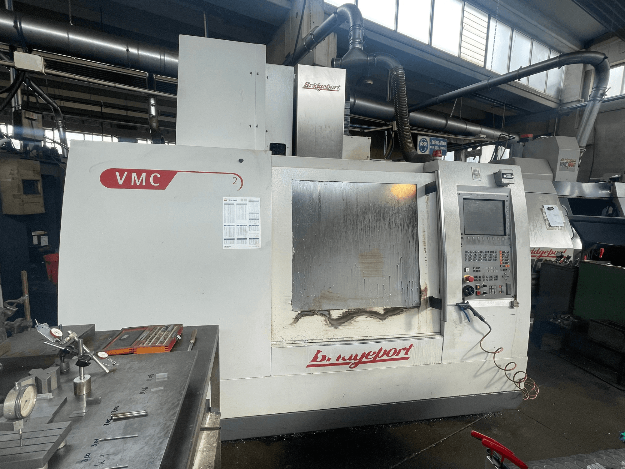 Вертикальный обрабатывающий центр Bridgeport VCM 800-30 в цехе с панелью управления и столом для инструментов. Вид спереди с видимыми деталями.