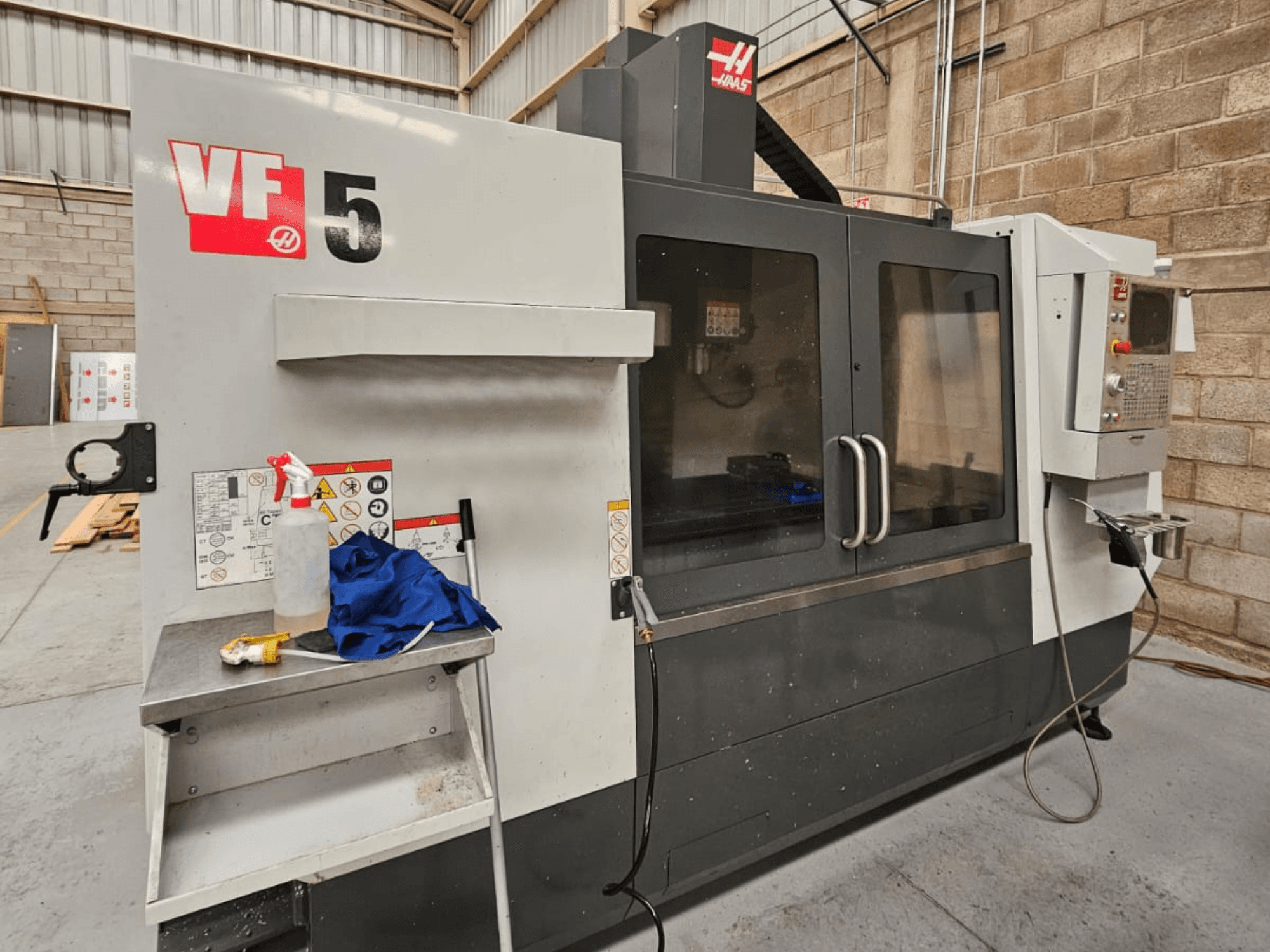 Фрезерный станок с ЧПУ Haas VF-5 с видом спереди, с панелью управления, бутылкой с охлаждающей жидкостью, синей тканью и инструкциями по технике безопасности.
