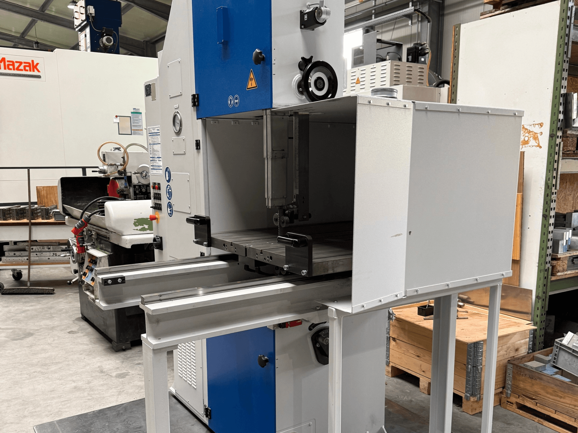 Промышленный станок Mazak с видом спереди, с синей панелью управления, металлическим столом и предупреждениями о безопасности.