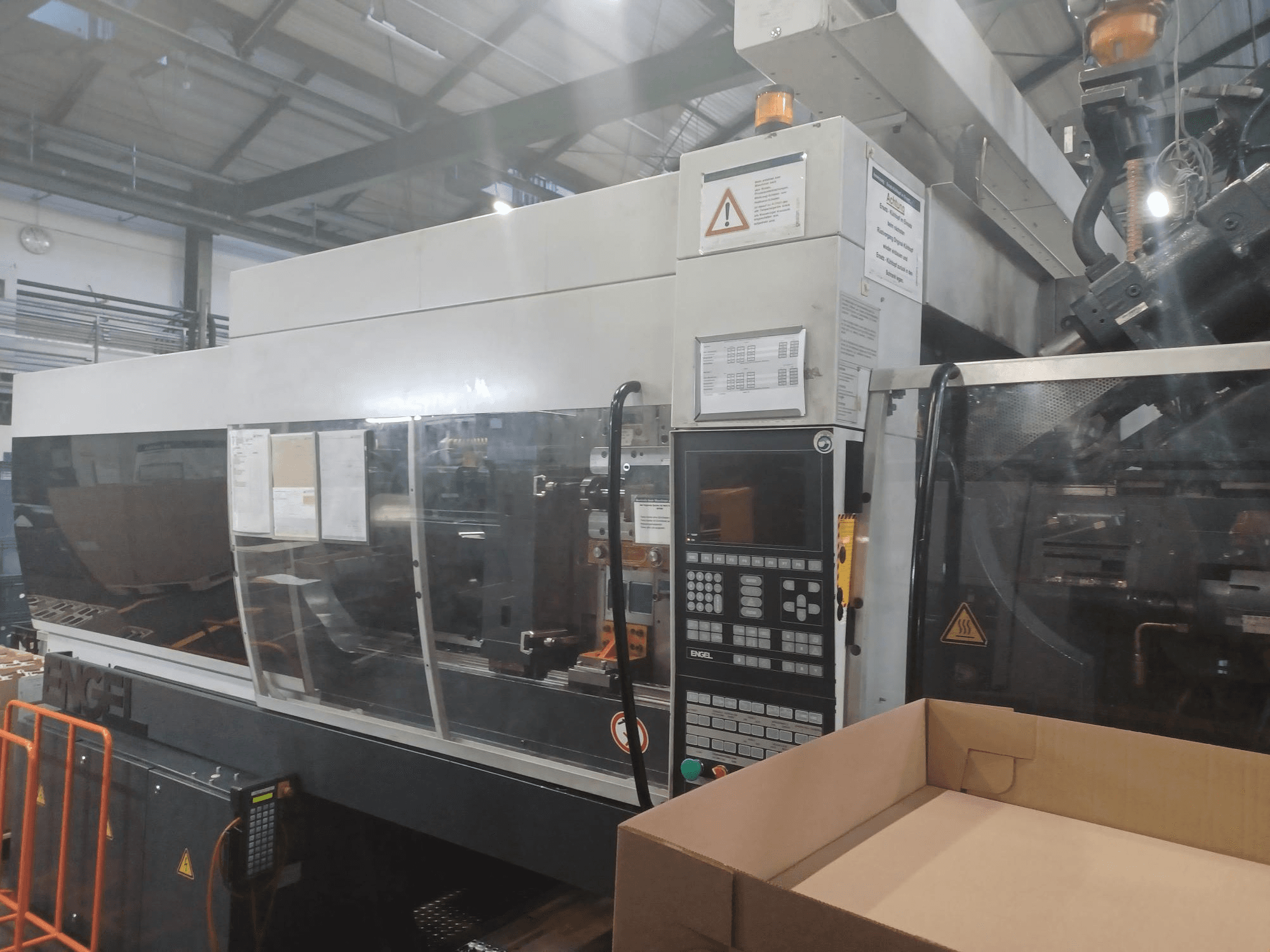 Термопластавтомат Engel, вид спереди, с панелью управления, наклейками безопасности и пустой коробкой, расположенной рядом.