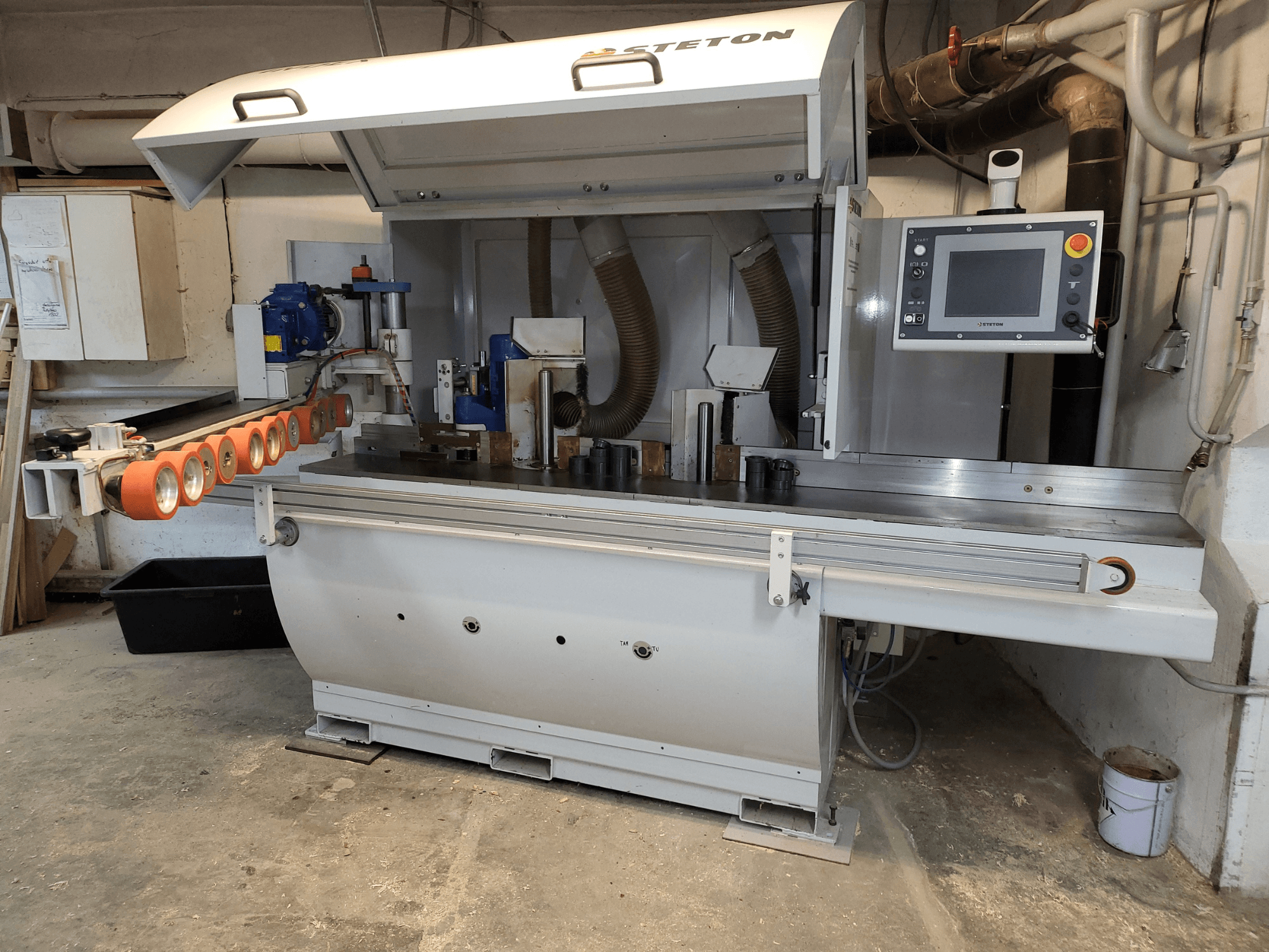 Вид спереди на деревообрабатывающий станок Stetton с панелью управления, подающими трубками и различными деревянными заготовками на столе.