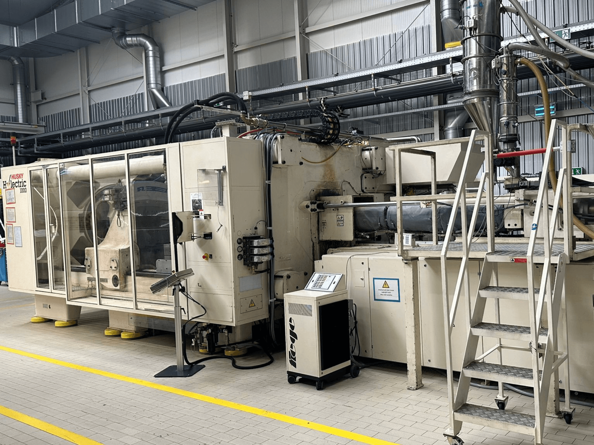 Вид спереди электрического термопластавтомата Husky Hylelectric H1000 RS95/80 в заводских условиях, с панелью управления и ступеньками.