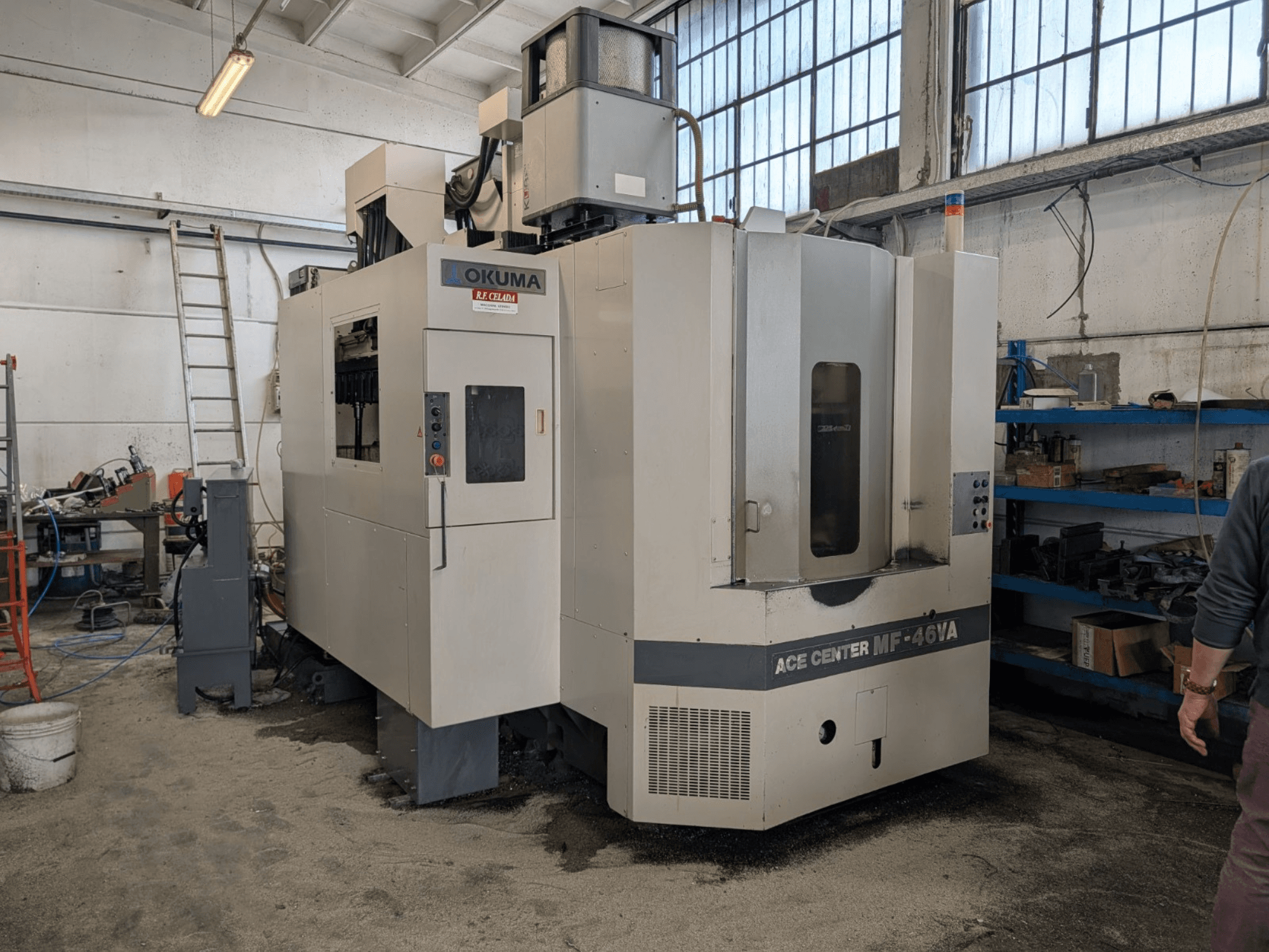 Вертикальный обрабатывающий центр Okuma ACE CENTER MF-46VA в мастерской, вид спереди с панелью управления и окружающим оборудованием.