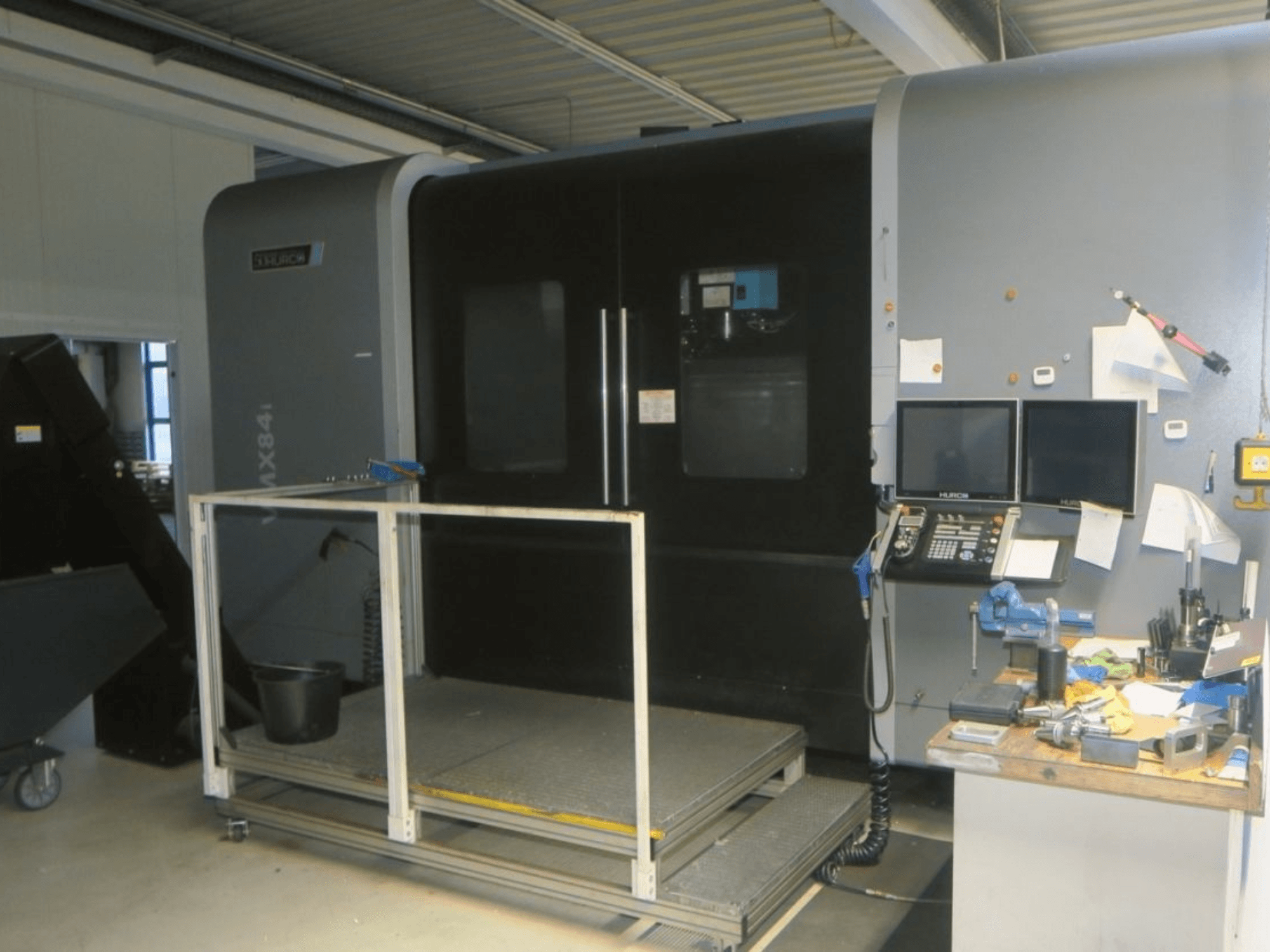 Станок с ЧПУ Hurco MX84 с видом спереди, показывает два монитора, панель управления и рабочую зону с различными инструментами.