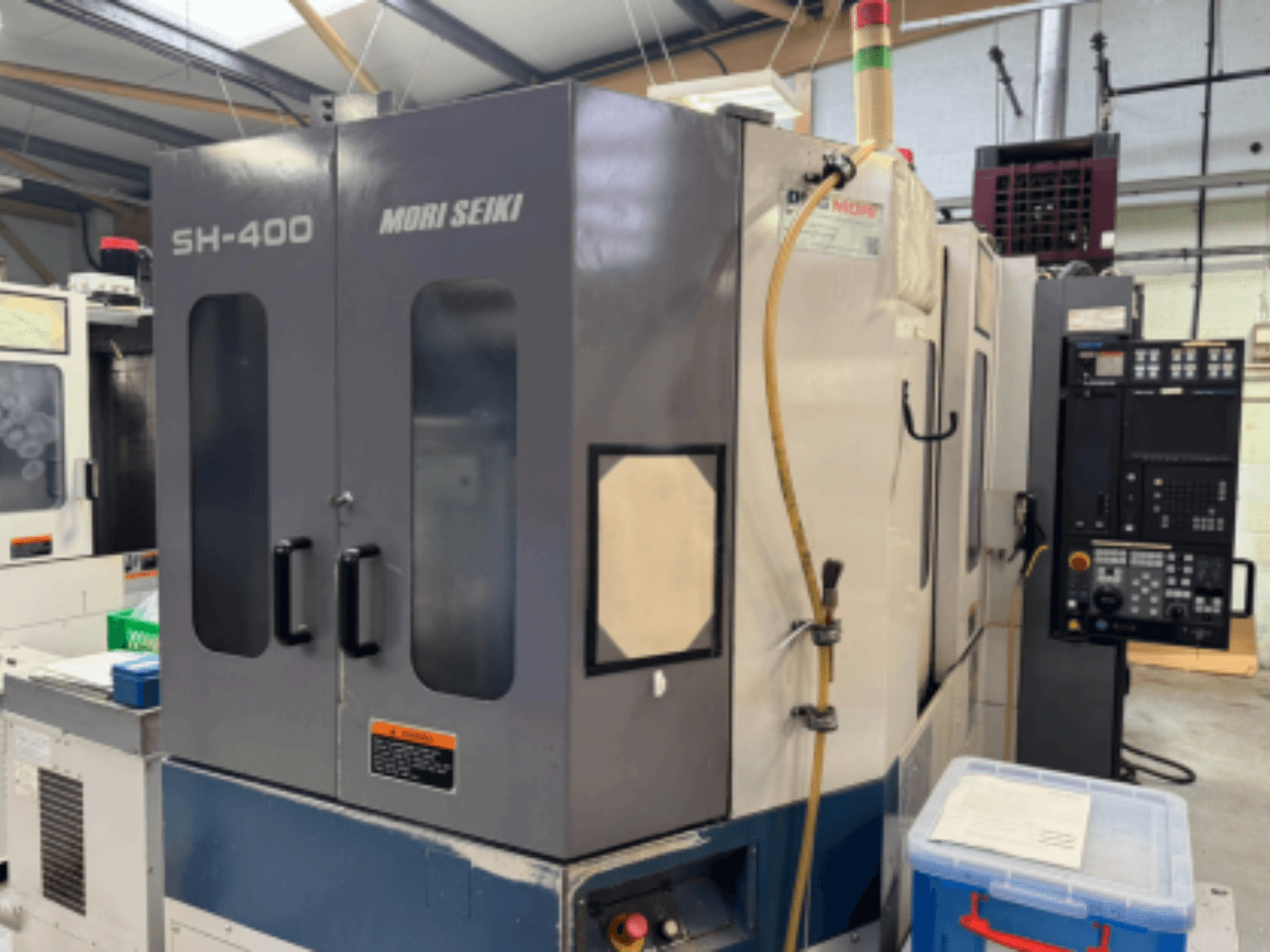 Станок с ЧПУ Mori Seiki SH-400, вид спереди, с двумя дверцами, панелью управления и шлангом для крепления. Синий контейнер для хранения рядом.