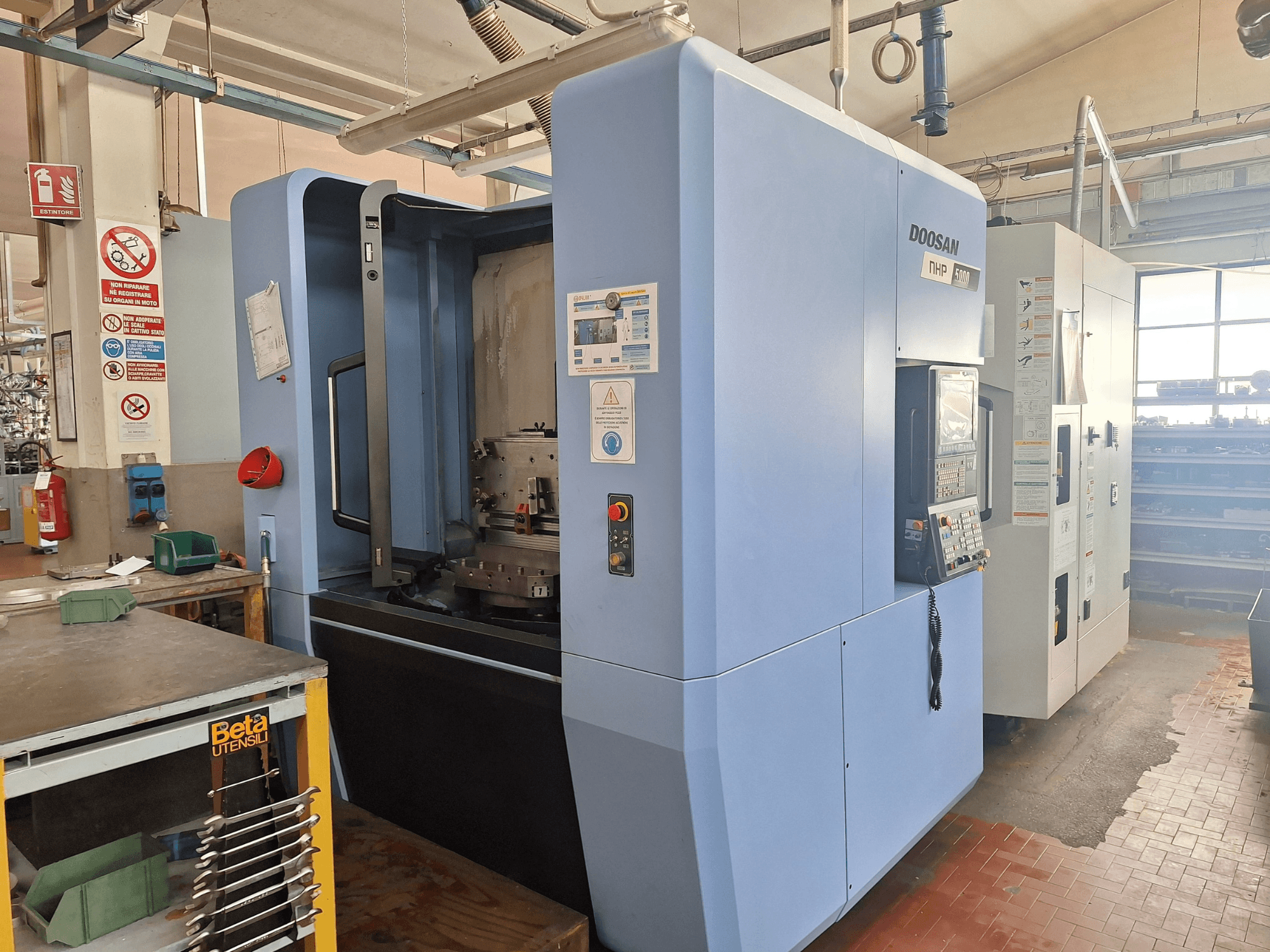 Горизонтальный обрабатывающий центр DOOSAN NHP 5000 в мастерской, вид сбоку, демонстрирующий панель управления и зону обработки.
