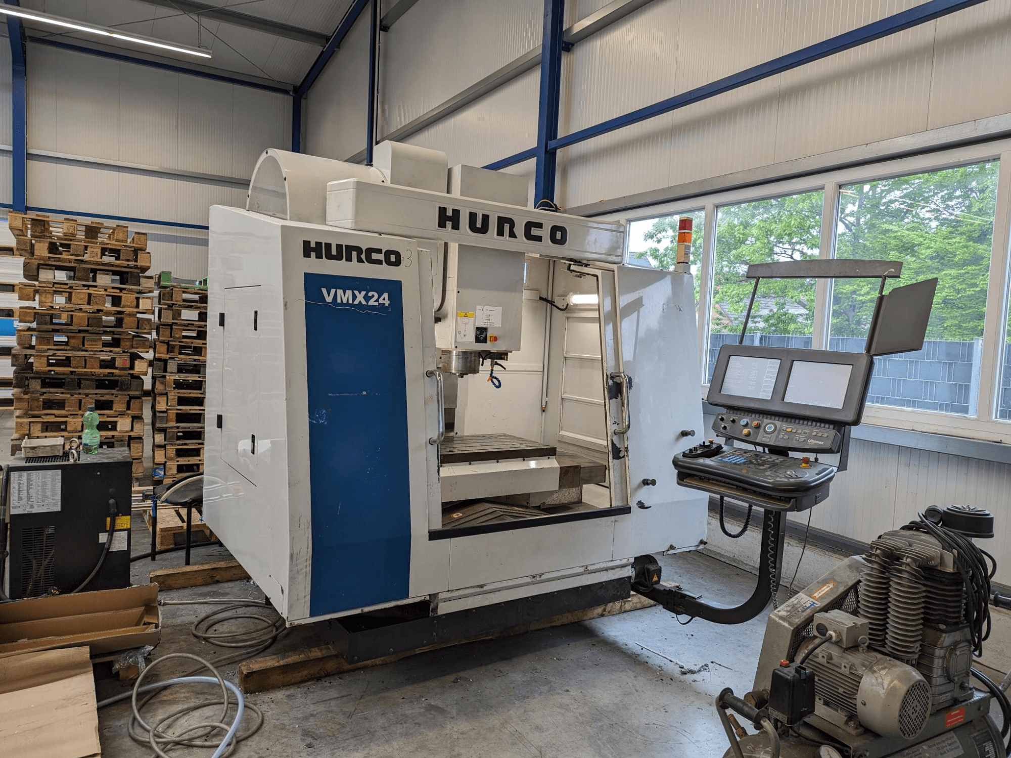 Фрезерный станок Hurco VMX24, вид спереди, с панелью управления и зоной инструментов; штабелированные паллеты на заднем плане; промышленное рабочее место.
