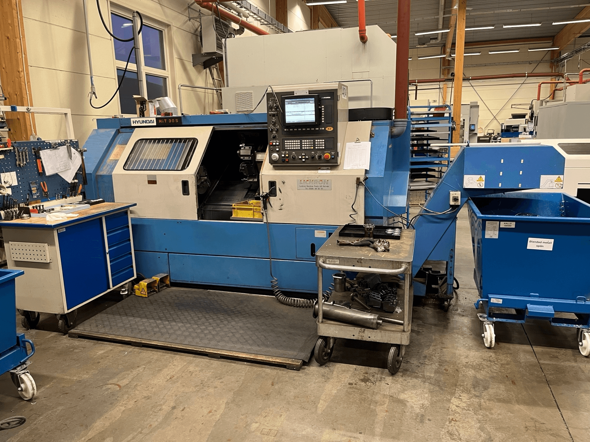 Горизонтальный токарный станок Hyundai HIT 30S, вид спереди, с панелью управления и инструментами на соседней тележке в мастерской.
