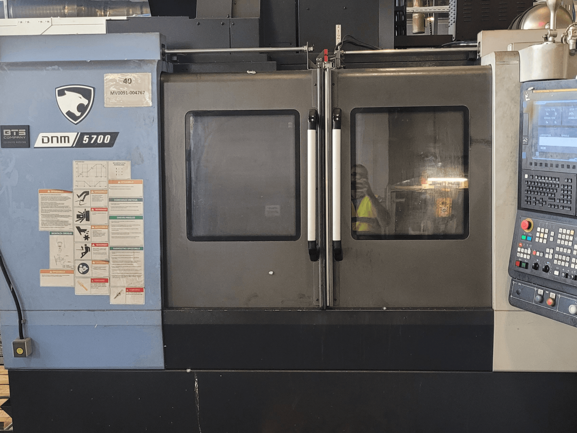 Вертикальный обрабатывающий центр DOOSAN DNM 5700, вид спереди: с панелью управления, предупреждающими надписями и слегка приоткрытыми дверцами.