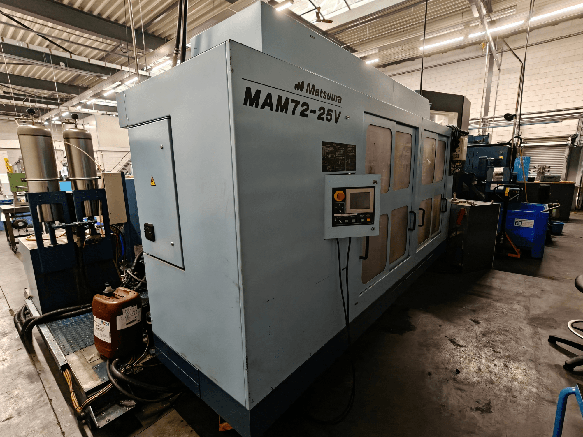Станок с ЧПУ Matsuura MAM72-25V, вид сбоку, панель управления, два бака с охлаждающей жидкостью и рабочее место в промышленной обстановке.