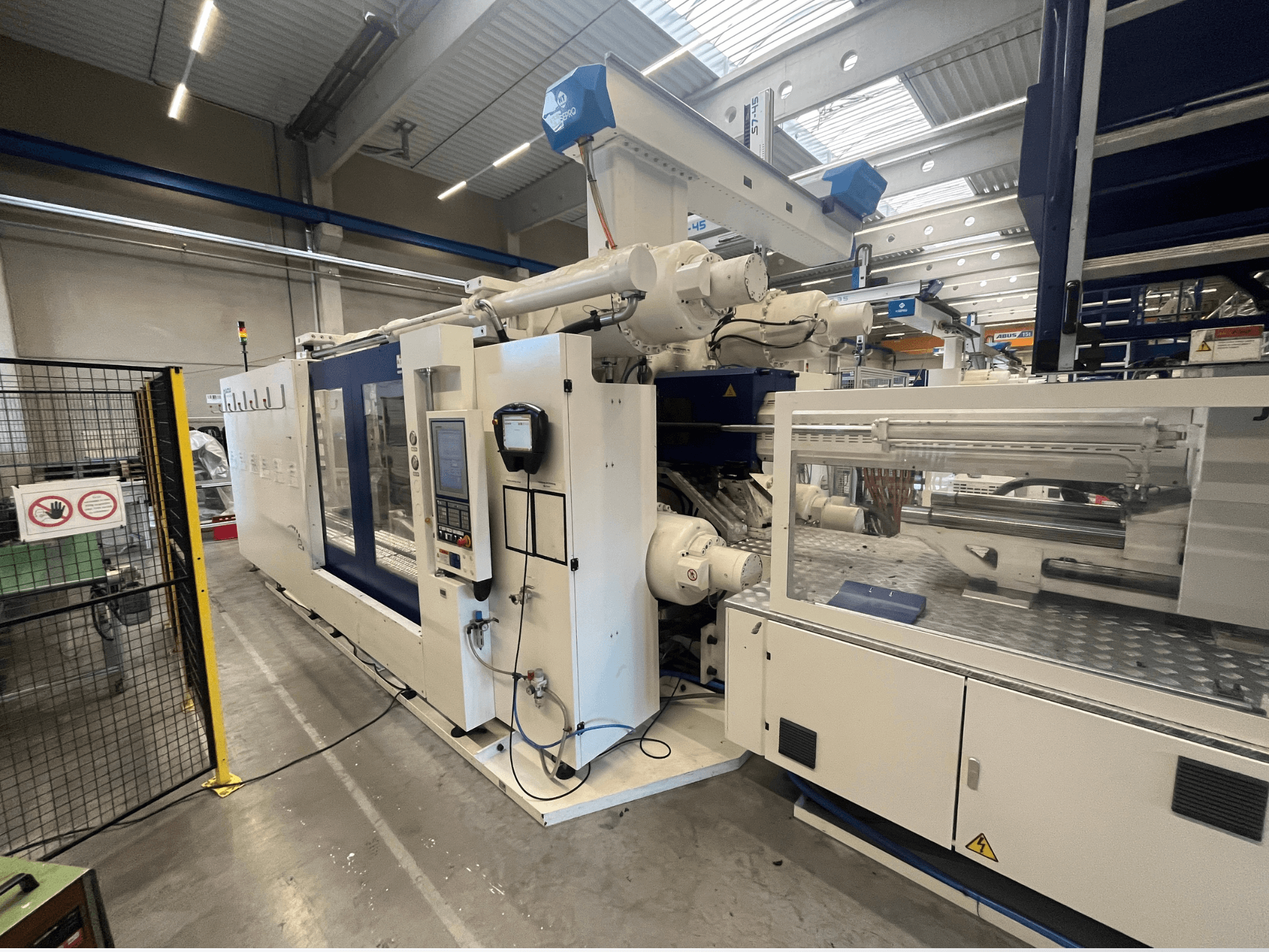 Машина для литья под давлением белого и синего цвета, вид спереди, с панелью управления, роботизированной рукой и знаками безопасности.