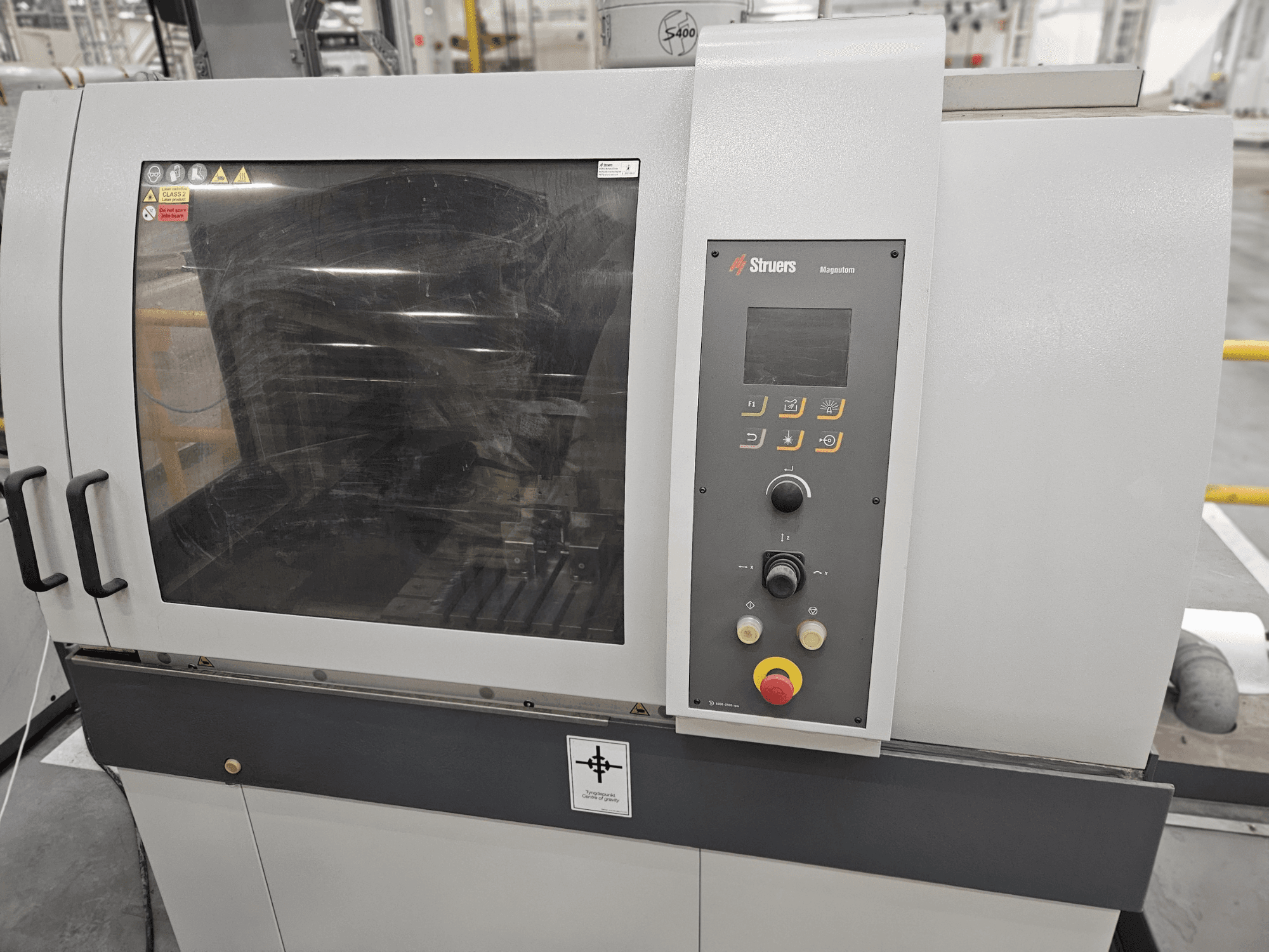 Вид спереди машины Struers Magpentor с панелью управления, кнопками и большим смотровым окном. Видны технические надписи.