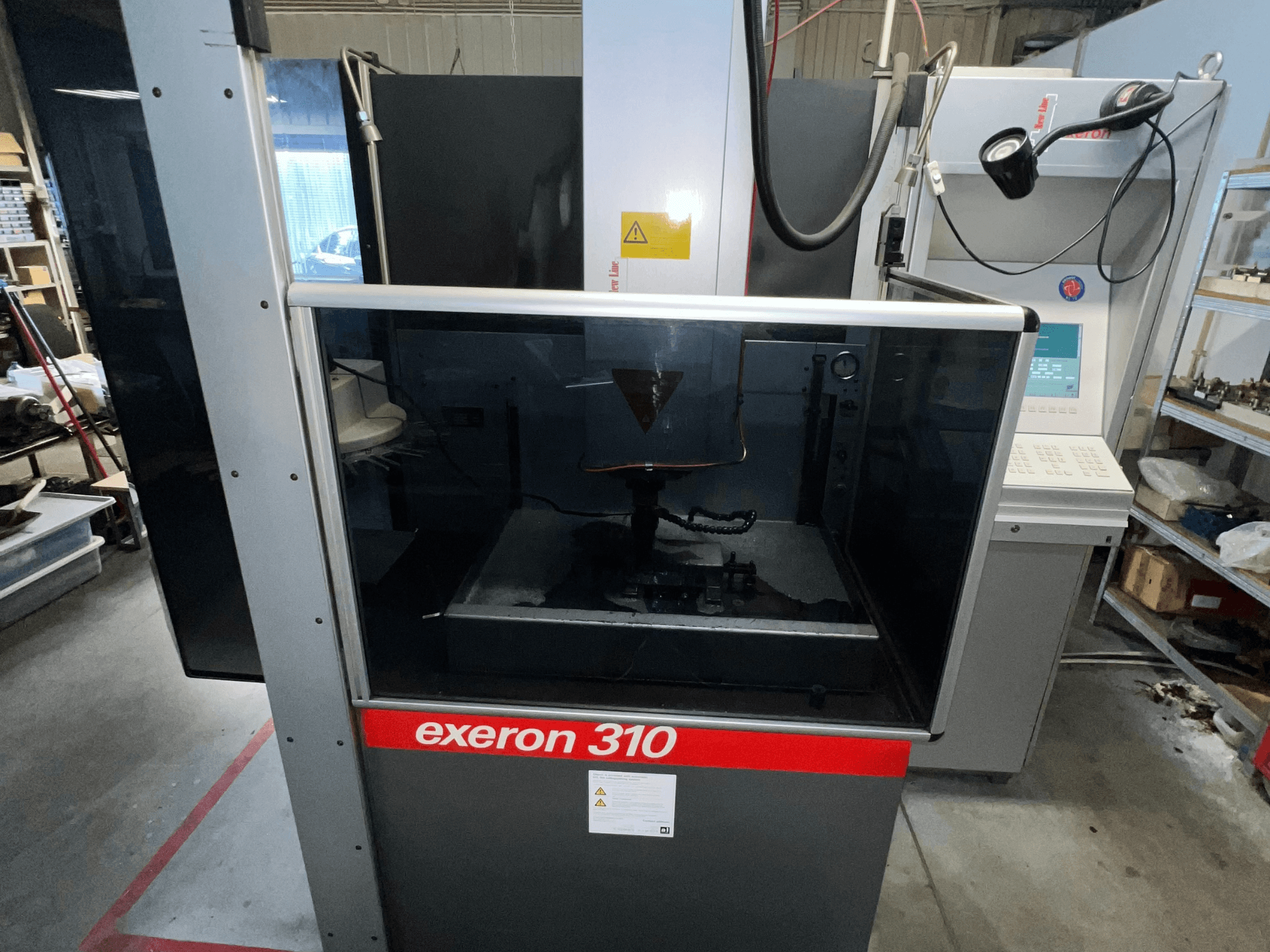 Вид спереди на машину Exeron 310, демонстрирующую стеклянную камеру и панель управления рядом со стеллажами для рабочих мест.