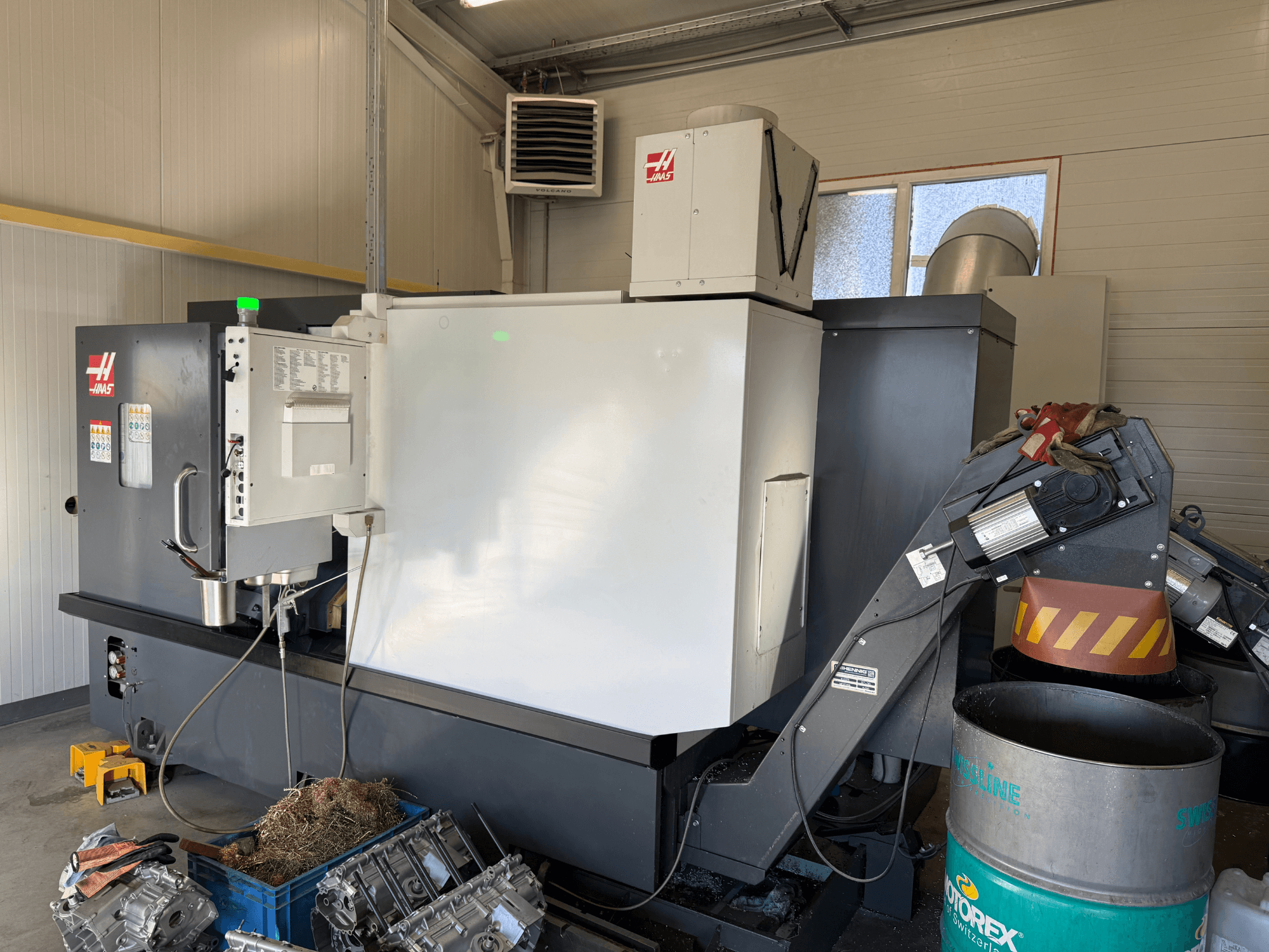 Станок с ЧПУ Haas с панелью управления, вид сбоку; видны трубопроводы охлаждающей жидкости и масла; металлические обрезки в синей корзине; промышленная обстановка.