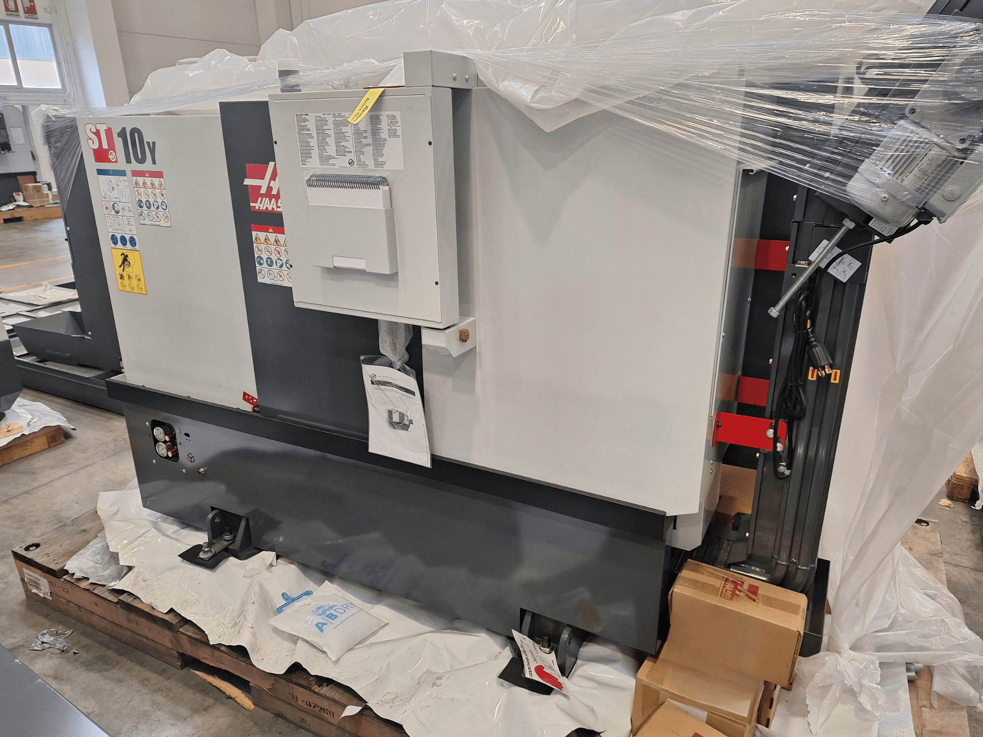 Горизонтальный токарный станок HAAS ST-10Y EU в виде сбоку, упакованный в пластиковую пленку, с видимой панелью управления и предупреждающими наклейками.