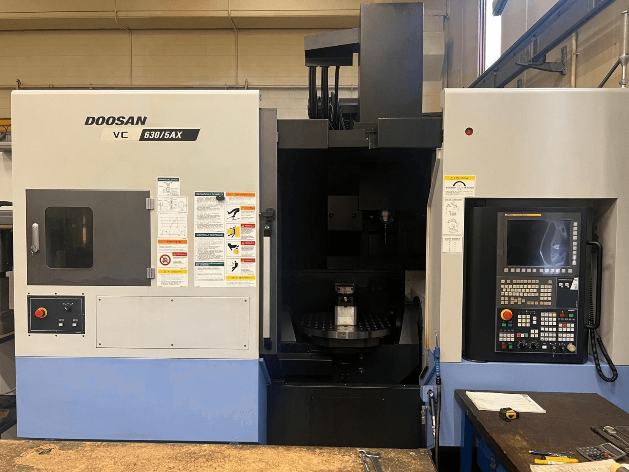 Вид спереди вертикального обрабатывающего центра DOOSAN VC 630/5AX, на котором видны панель управления и зона шпинделя с наклеенными предупреждающими надписями.