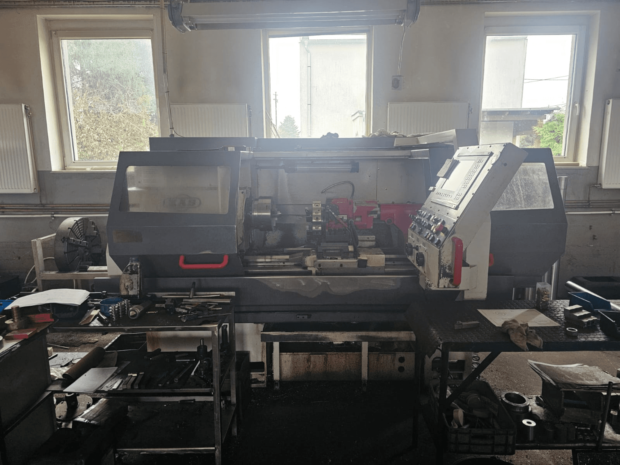 Токарный станок в мастерской, вид спереди, с панелью управления и инструментами на столах, в окружении окон и загроможденного рабочего пространства.