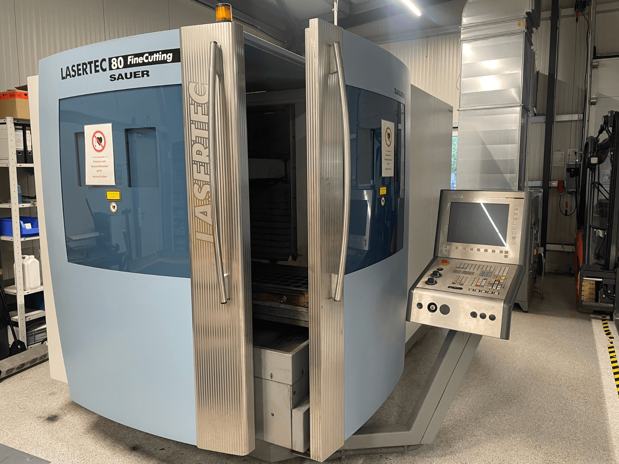 Вид спереди на станок для тонкой резки листового металла DMG Sauer Lasertec80 FineCutting с панелью управления, открытыми дверцами и видимыми знаками безопасности.